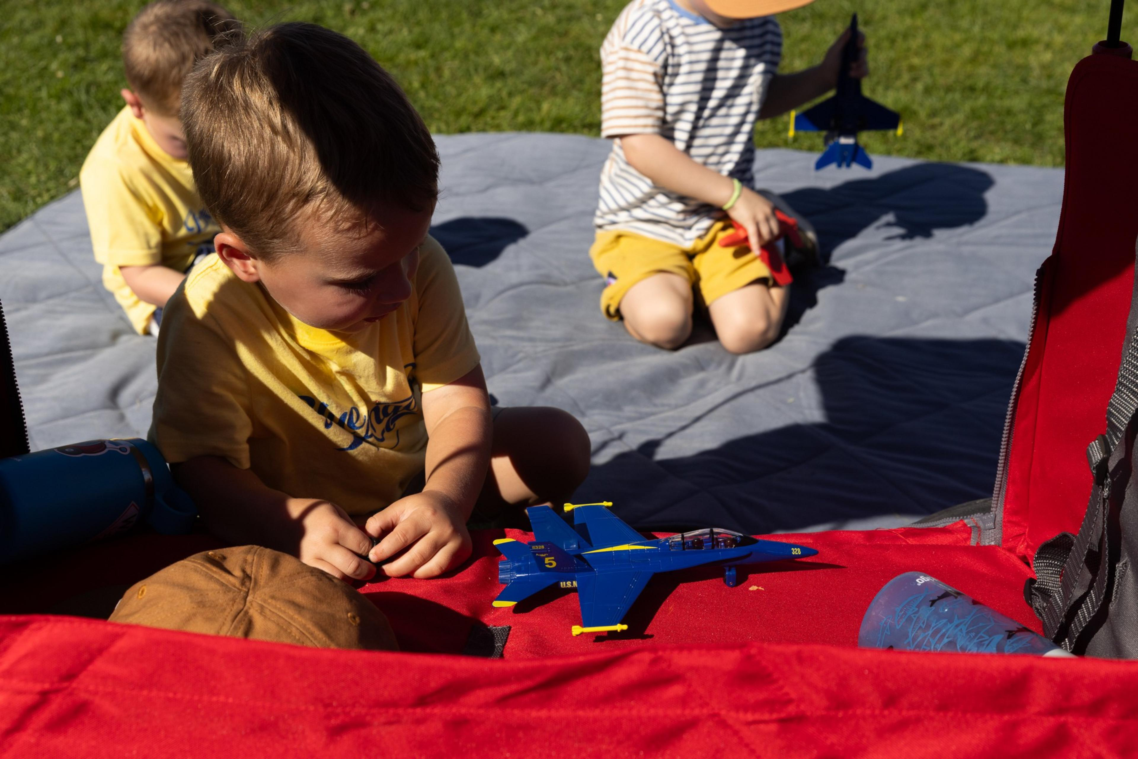 Three young boys play outdoors on a gray blanket; one boy in yellow focuses on a blue toy airplane on a red surface nearby.