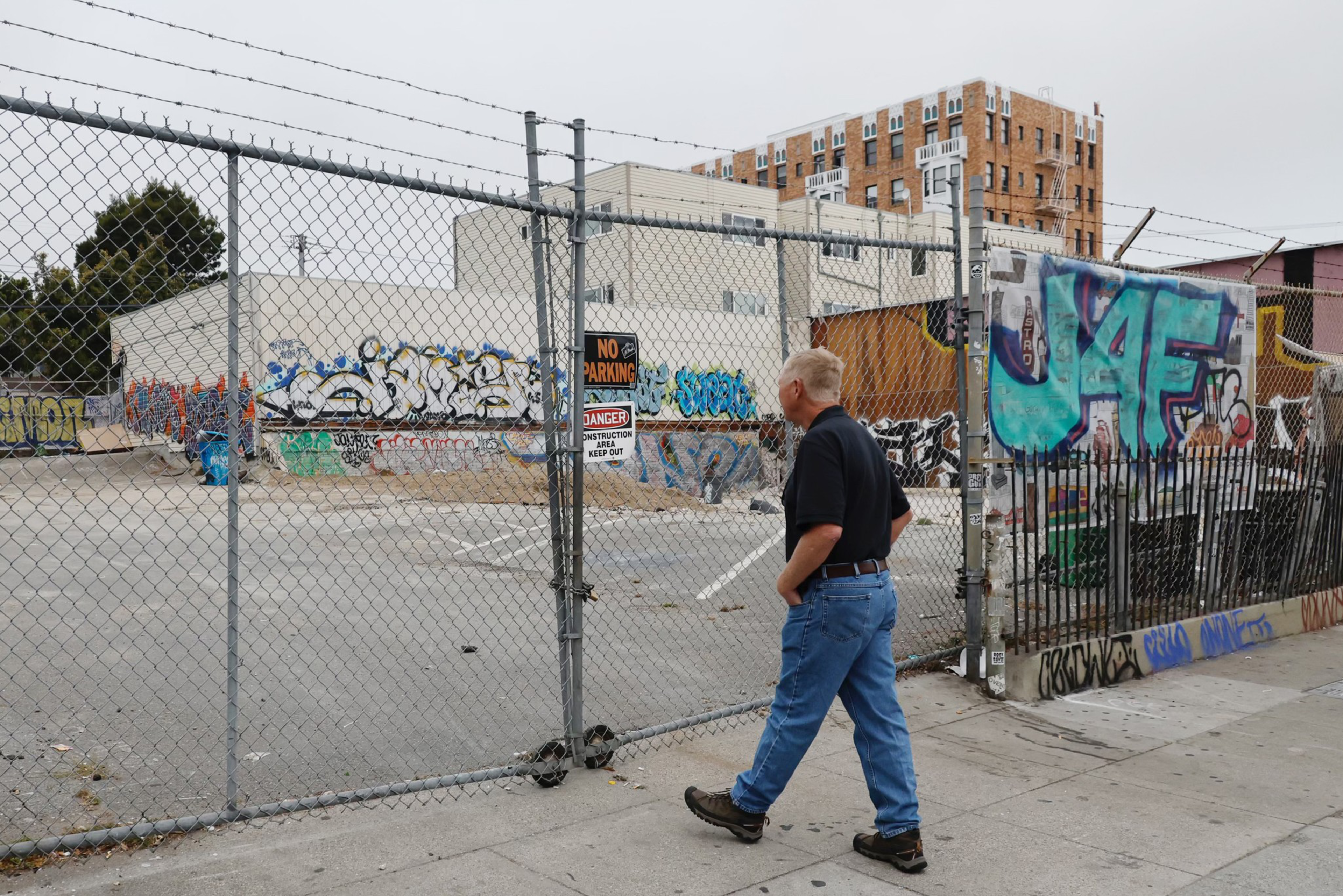 A man in a black shirt and blue jeans walks past a fenced, graffiti-covered empty lot with various buildings in the background under a gray sky.