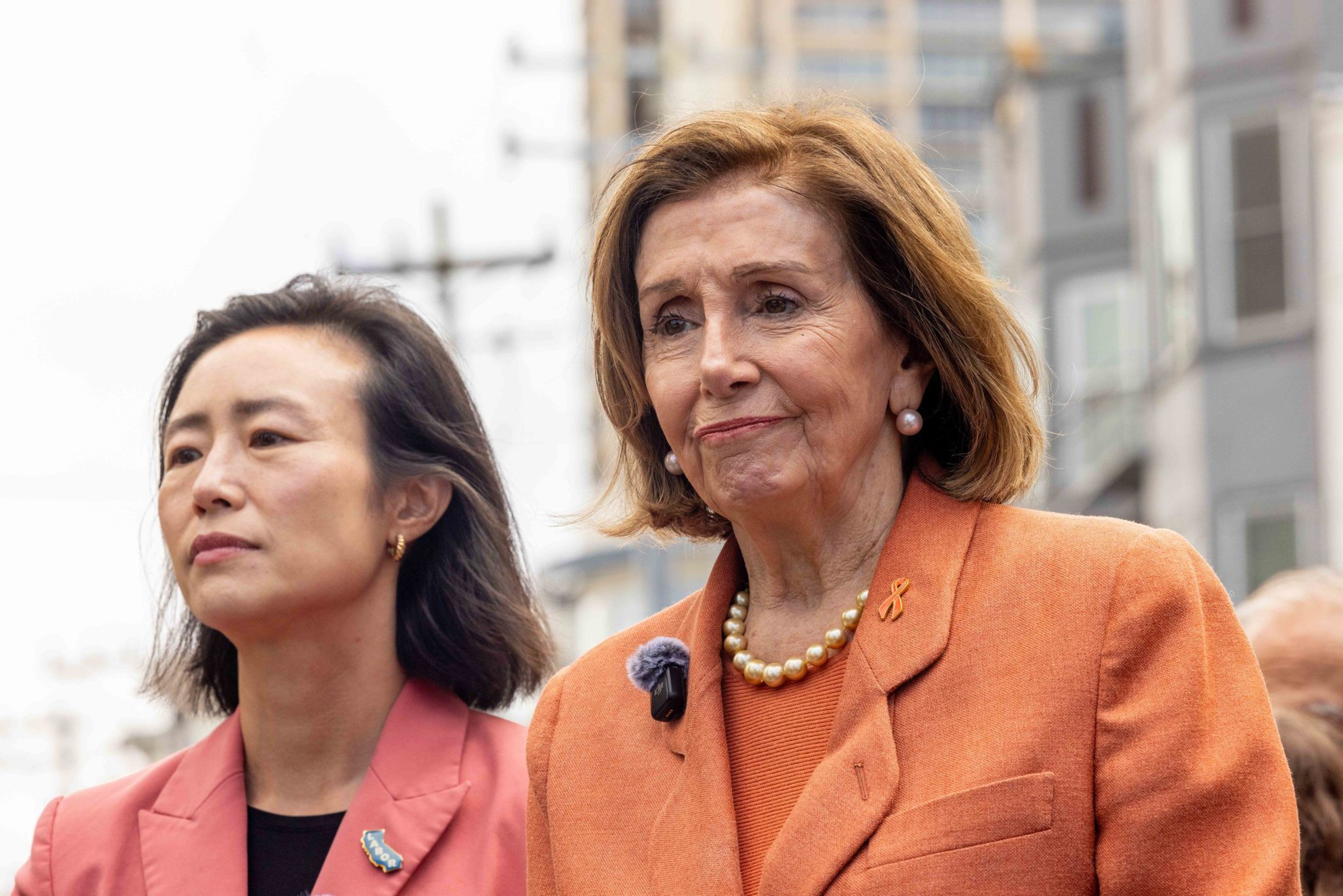 Two women stand side by side outdoors, both wearing orange blazers; one has a pearl necklace and an orange ribbon pin, appearing thoughtful.