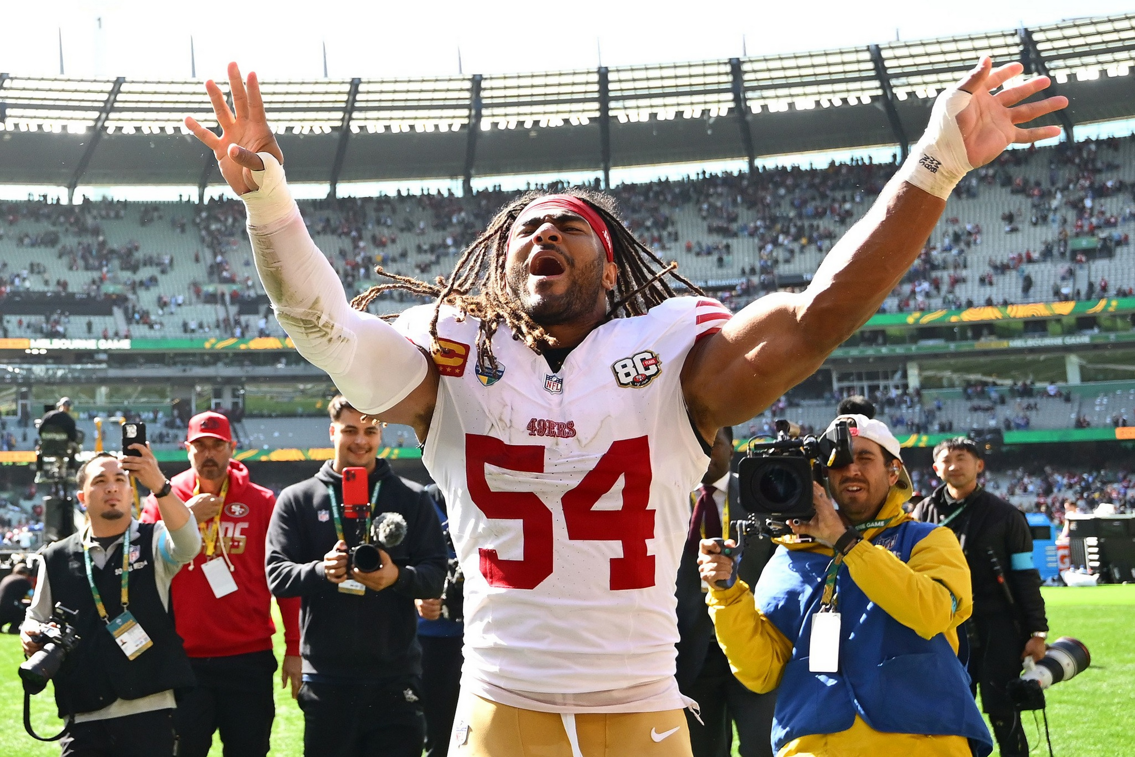 A football player wearing a white jersey with number 54 raises his arms celebrating on the field, surrounded by photographers capturing the moment.