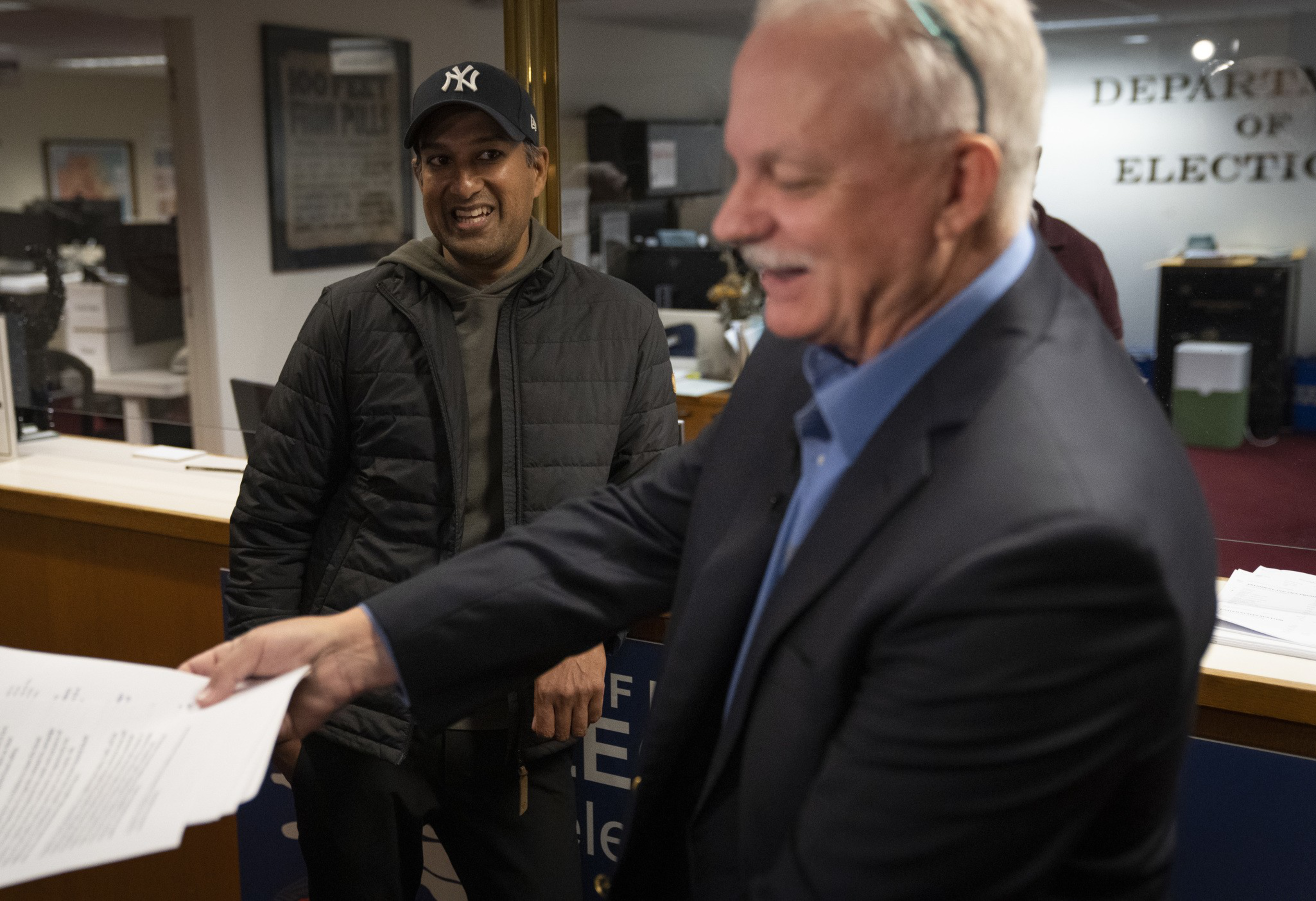 Two men are in an office setting. One, wearing a suit, is smiling while handing over papers. The other, in a casual jacket and cap, is smiling nearby.