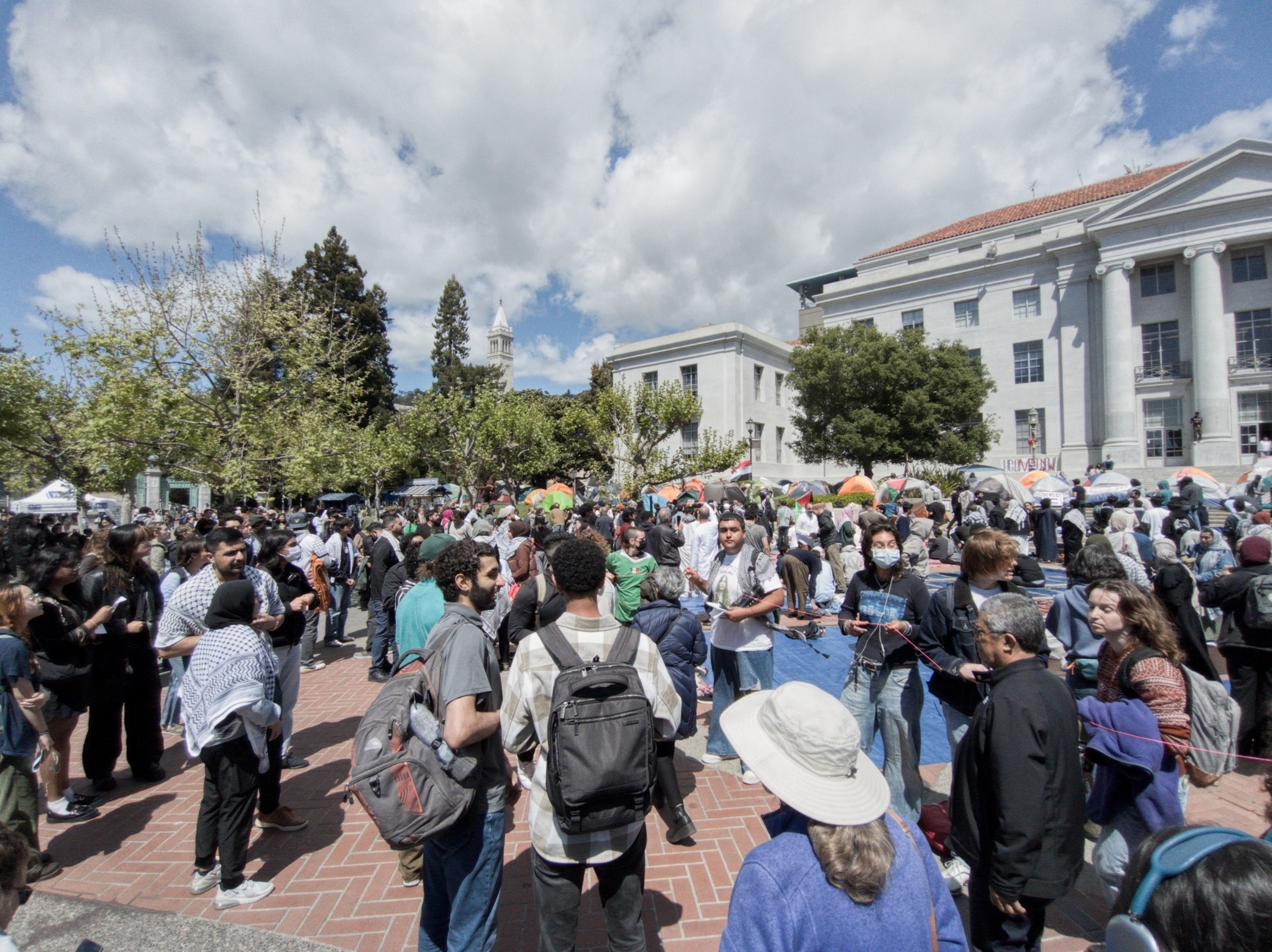A busy outdoor gathering with a diverse crowd in a public square, backed by a stately building and trees under a partly cloudy sky.