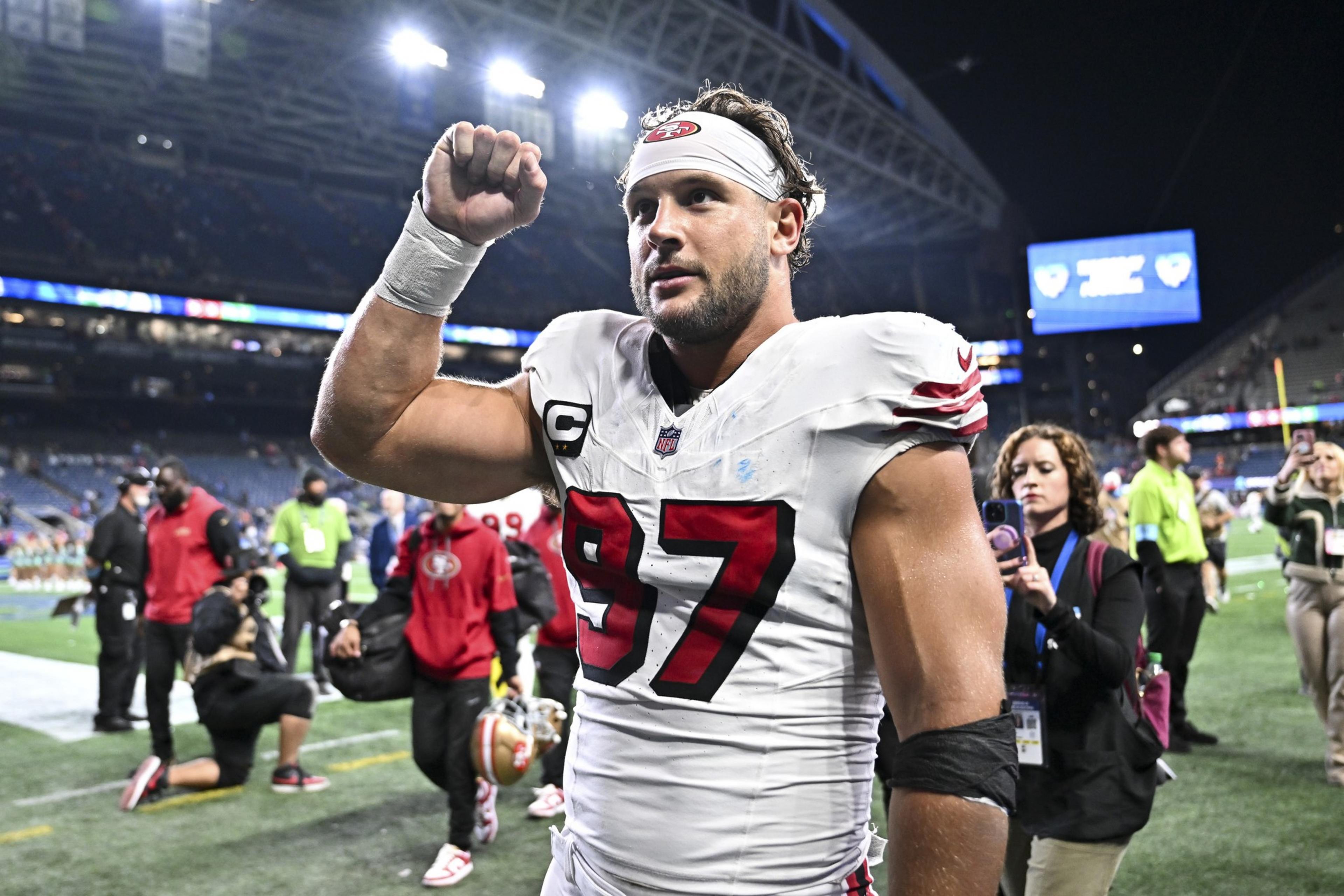 A football player in a white and red uniform with the number 97 is on a field. He raises his arm in a triumphant gesture, surrounded by people and stadium lights.