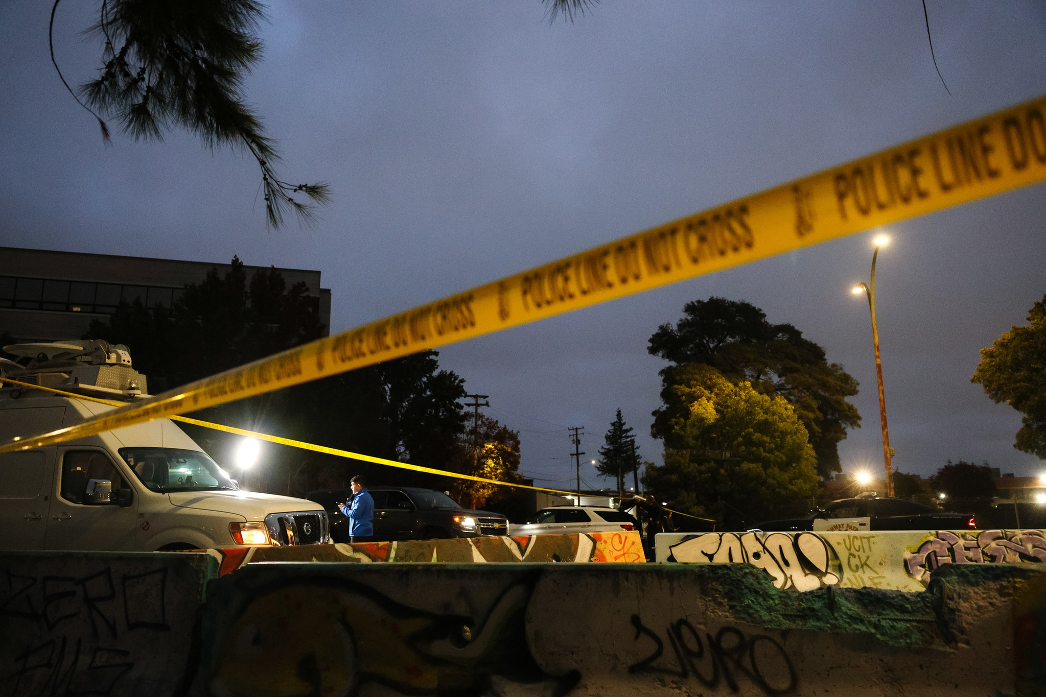 Yellow police tape stretches across a dimly lit outdoor scene at dusk, with a news van, a person in blue, and graffiti-covered barriers visible.