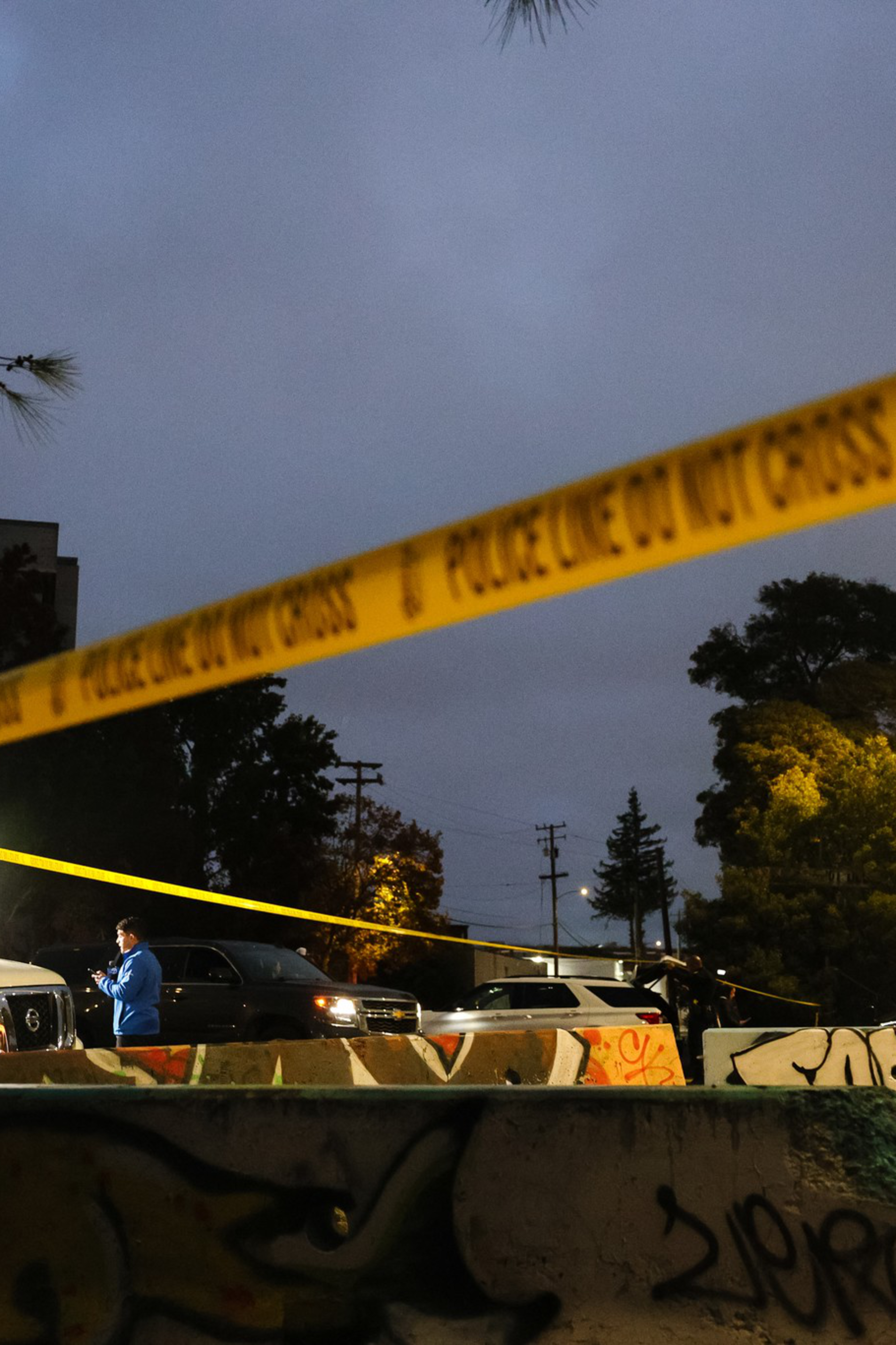 Yellow police tape stretches across a dimly lit outdoor scene at dusk, with a news van, a person in blue, and graffiti-covered barriers visible.