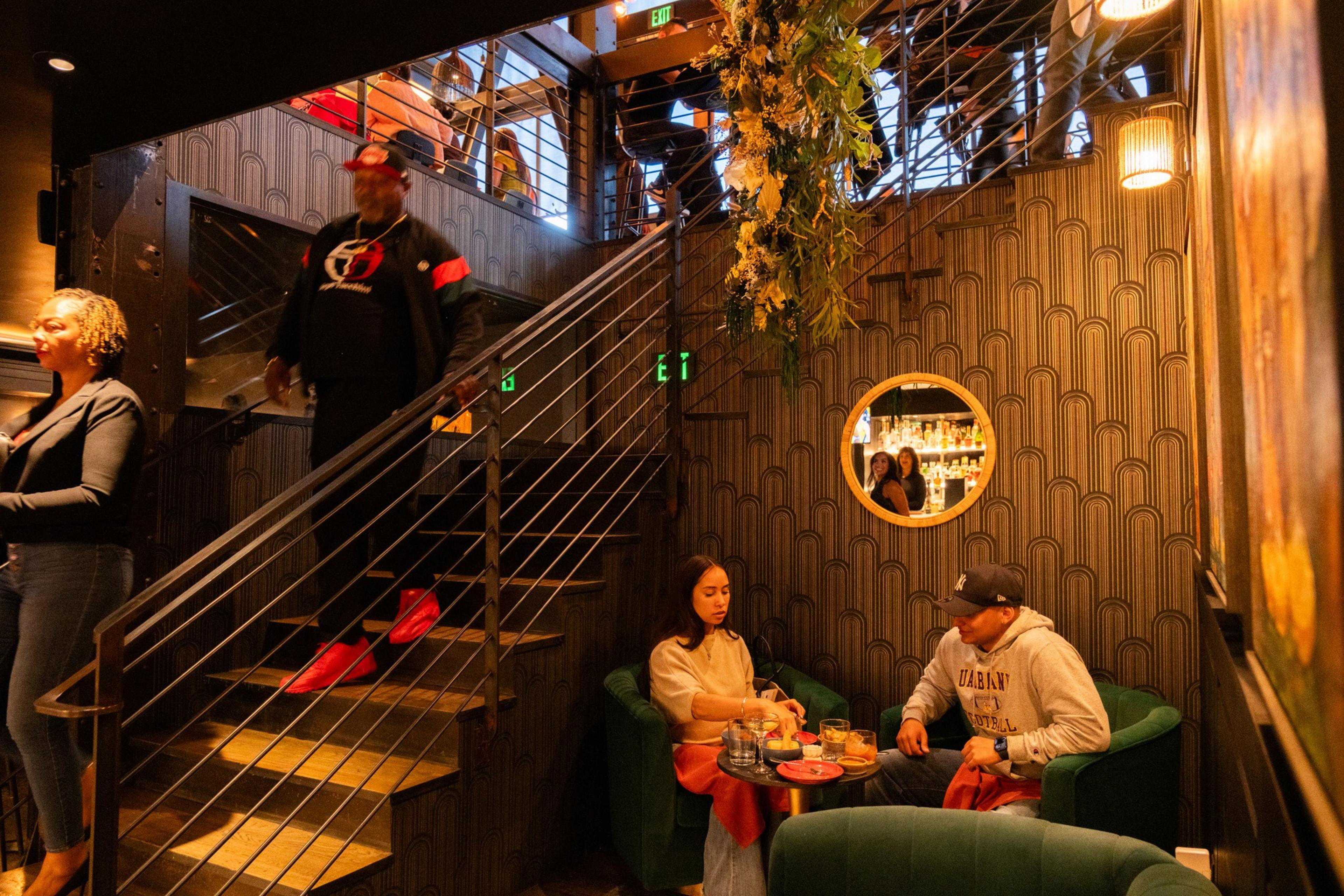 A stylish room features a staircase, patterned wallpaper, and a round mirror. Two people sit at a table with drinks, while others descend the stairs.