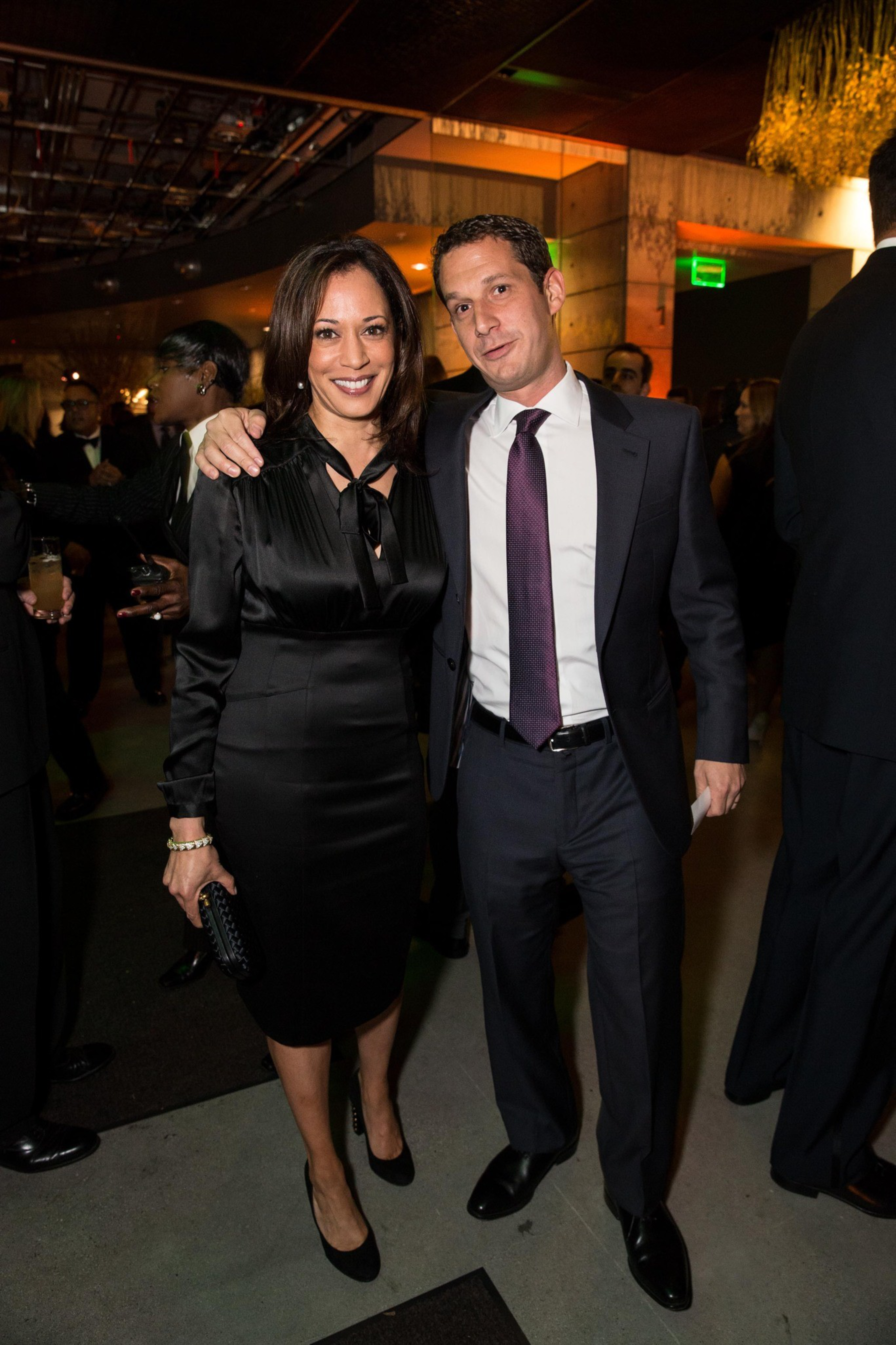 A woman in a black dress and a man in a suit with a purple tie stand smiling at an indoor event, surrounded by other people.