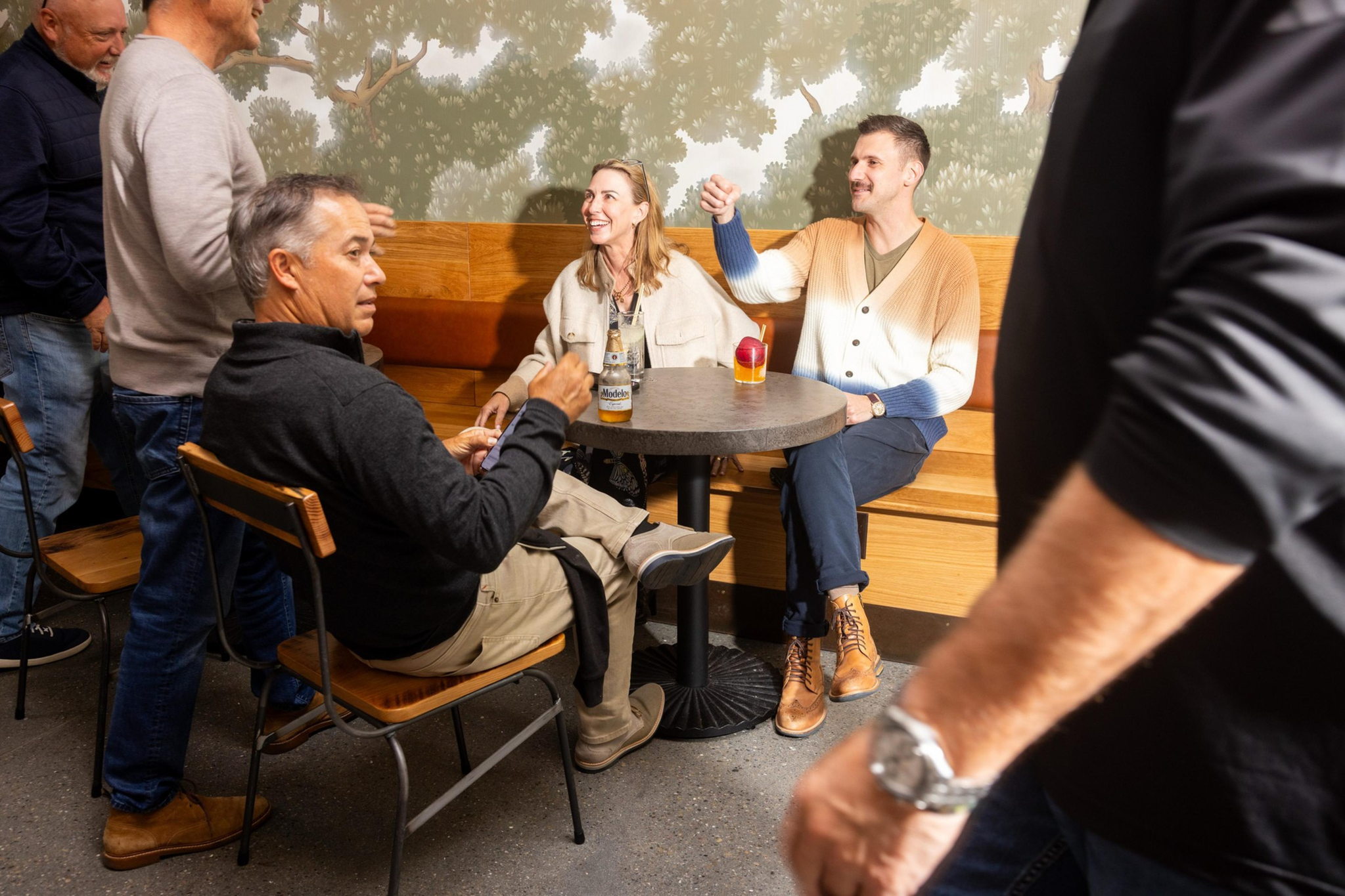 Several people are gathered in a casual indoor setting. Three are seated at a round table, with drinks, while others stand nearby, all engaged in conversation.
