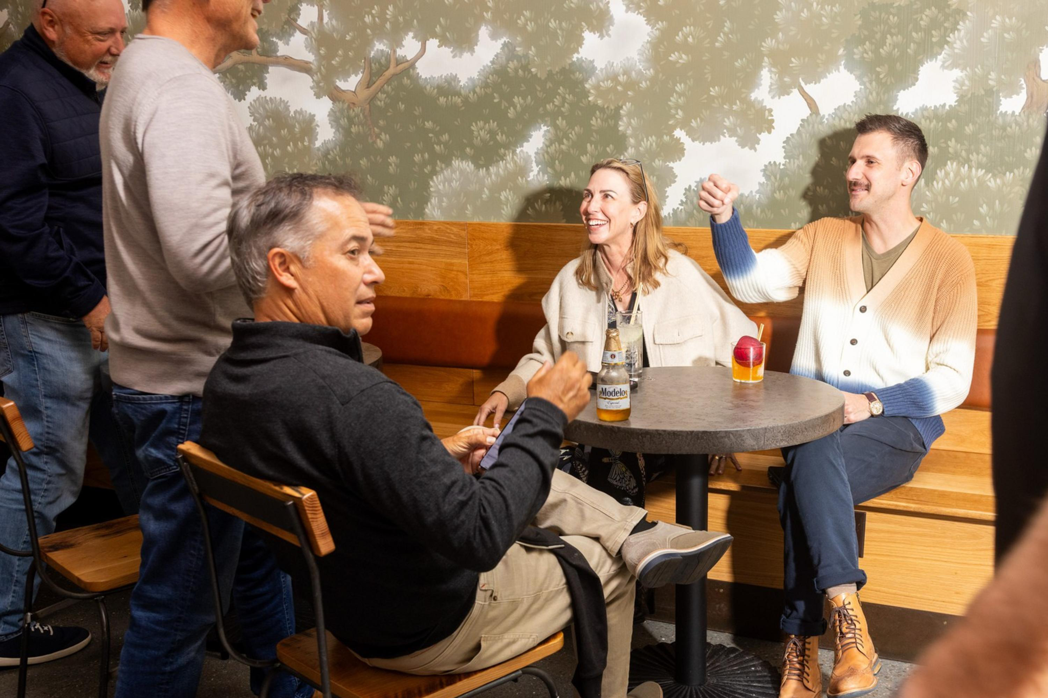 Several people are gathered in a casual indoor setting. Three are seated at a round table, with drinks, while others stand nearby, all engaged in conversation.