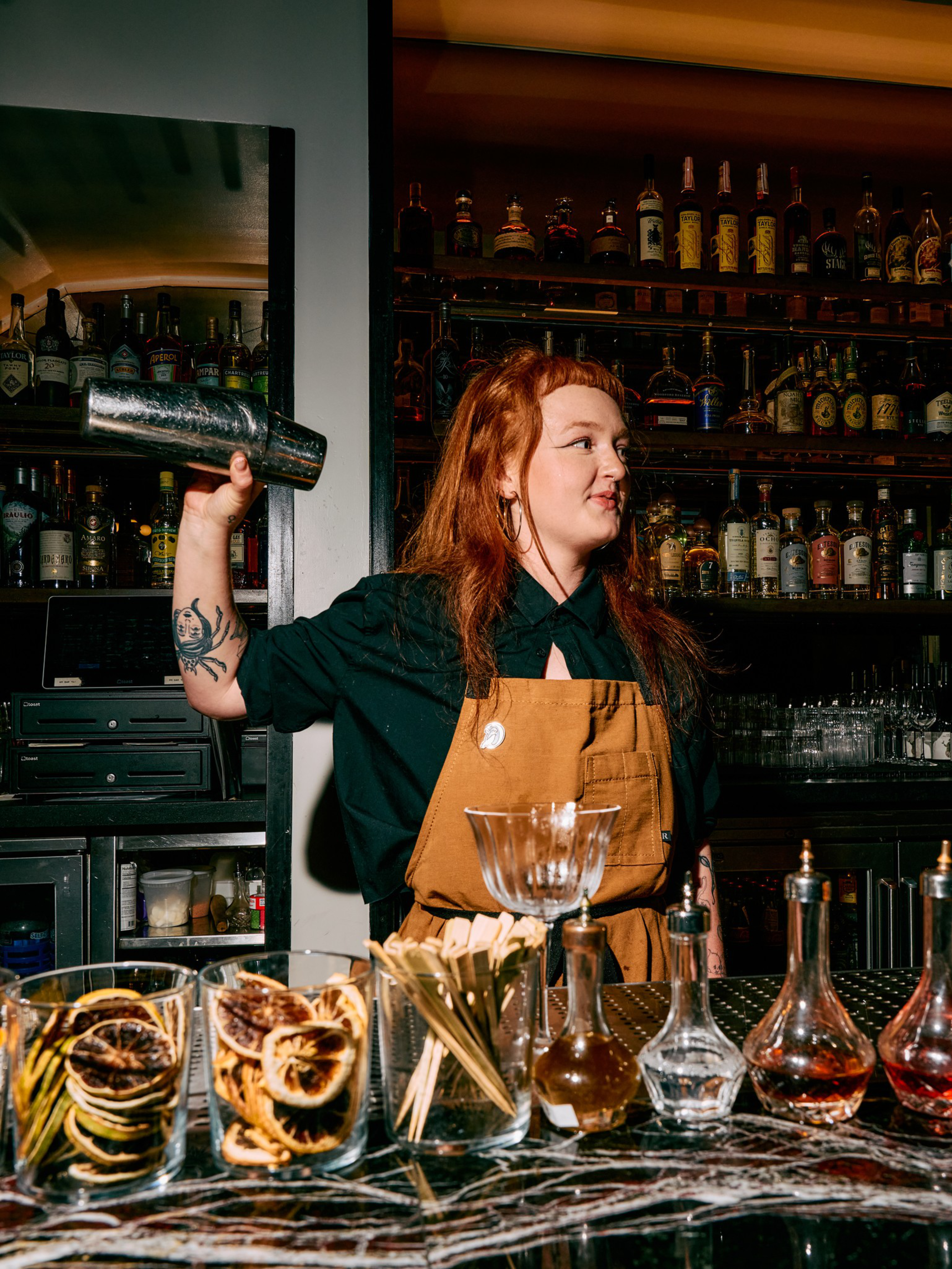A bartender with red hair and a tattoo shakes a cocktail mixer behind a bar filled with dried citrus, bottles, and glassware.