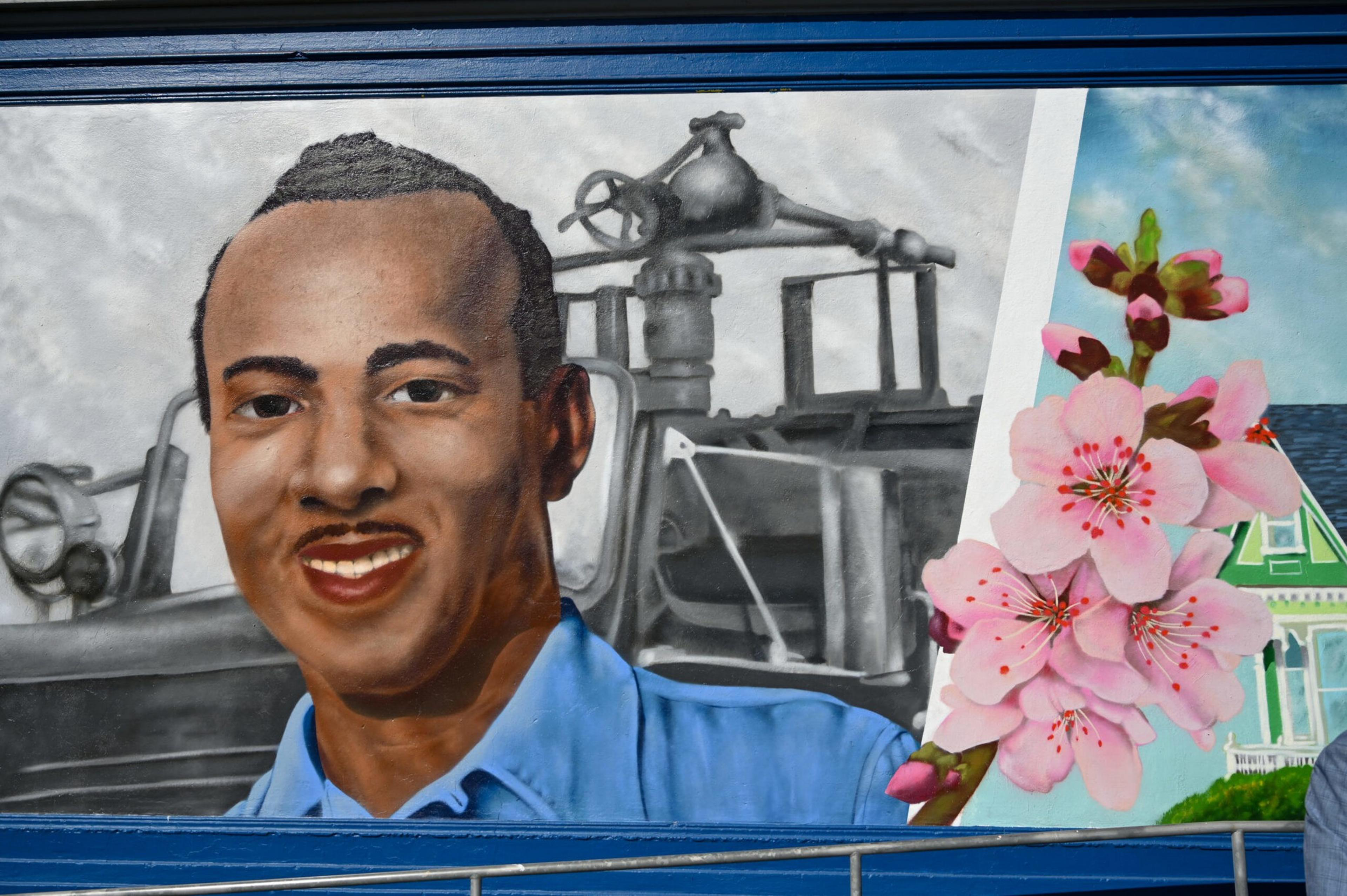 A mural features a smiling man in a blue shirt, an older industrial vehicle in grayscale behind him, and pink cherry blossoms with a green house on the right.