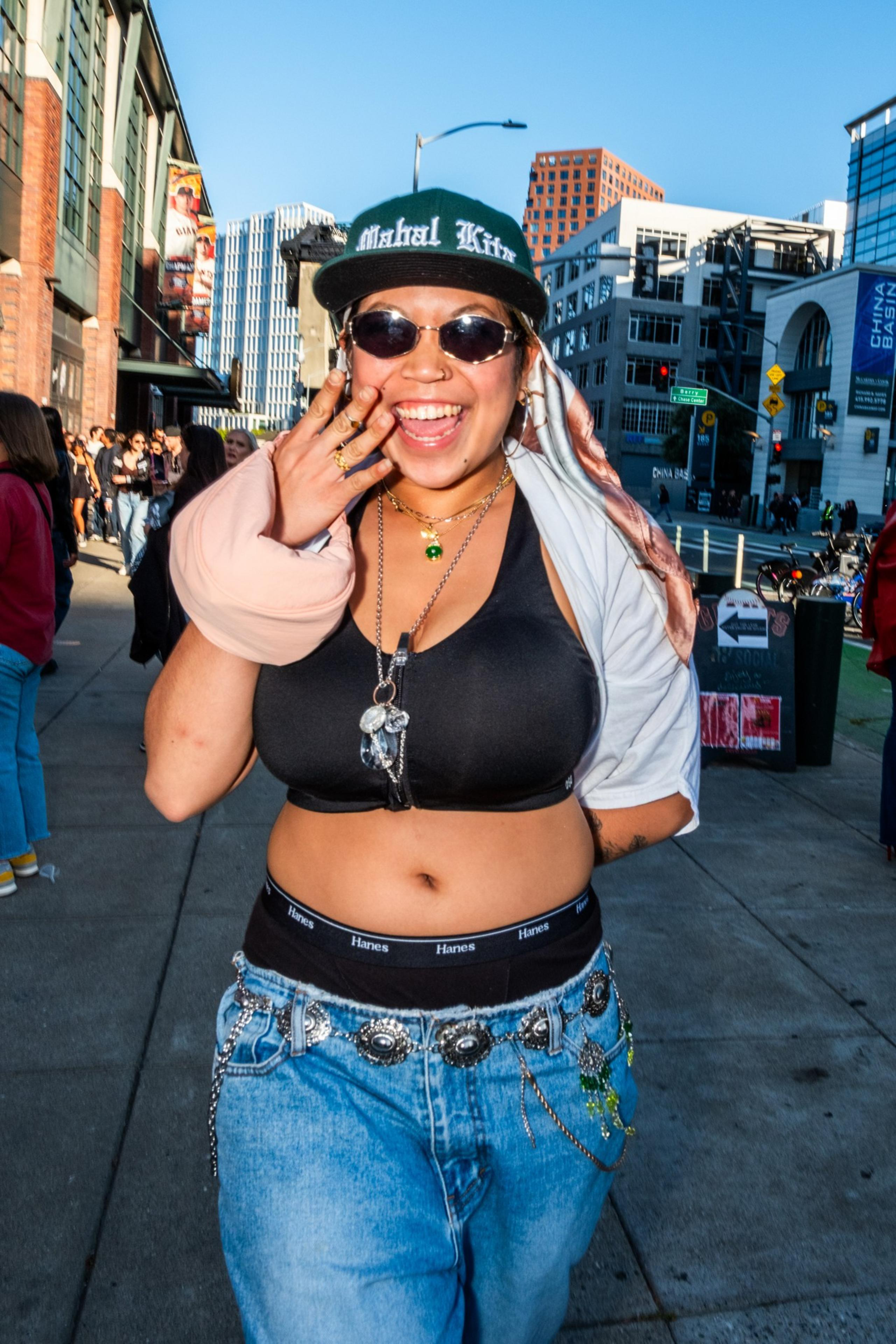 A smiling person wears sunglasses, a cap, a black crop top, and jeans with chains. They are outdoors in a lively urban area, posing energetically for the photo.