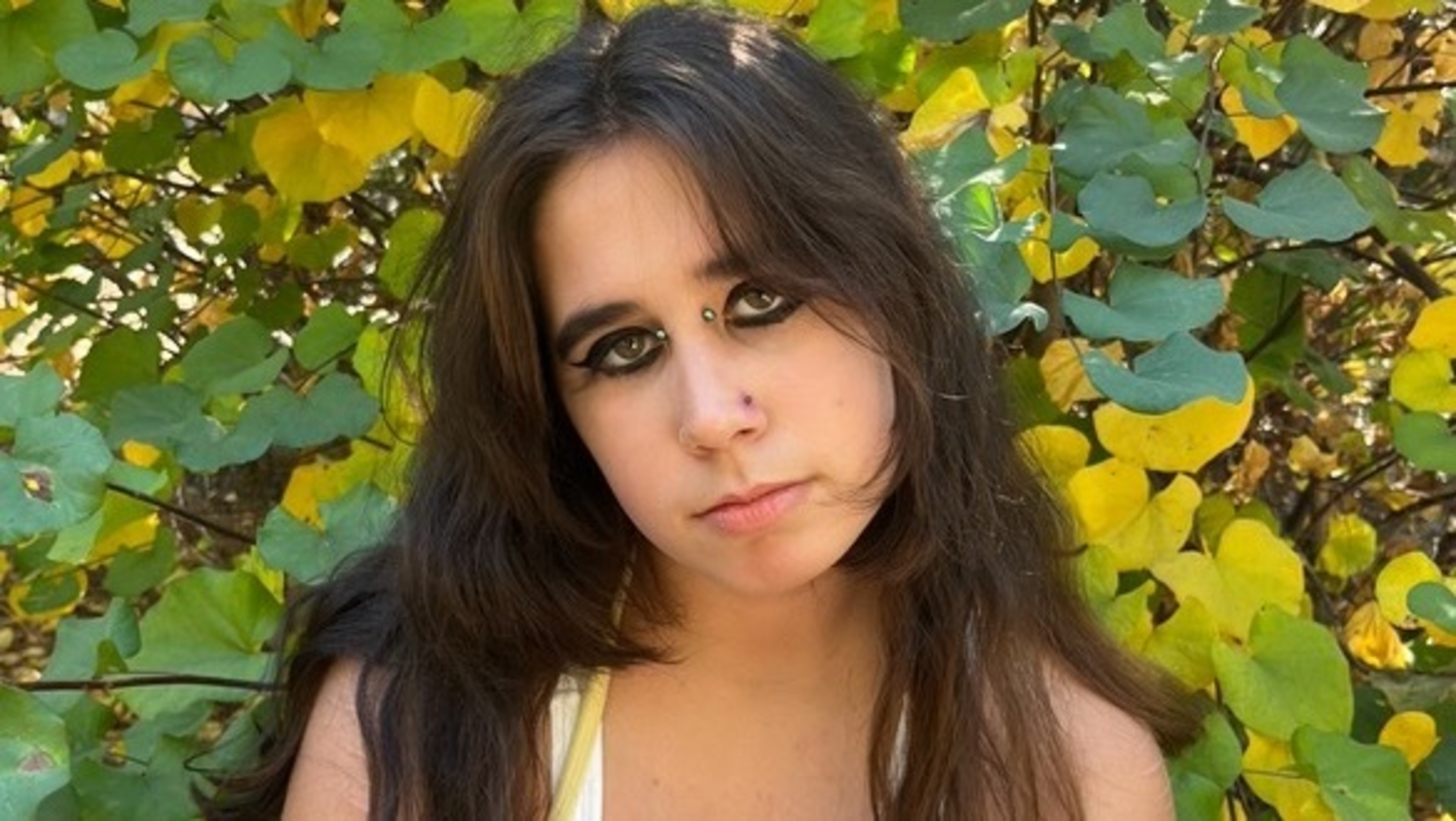 A dark haired young woman stands in front of green and yellow tree leaves, and looks directly at the viewer.