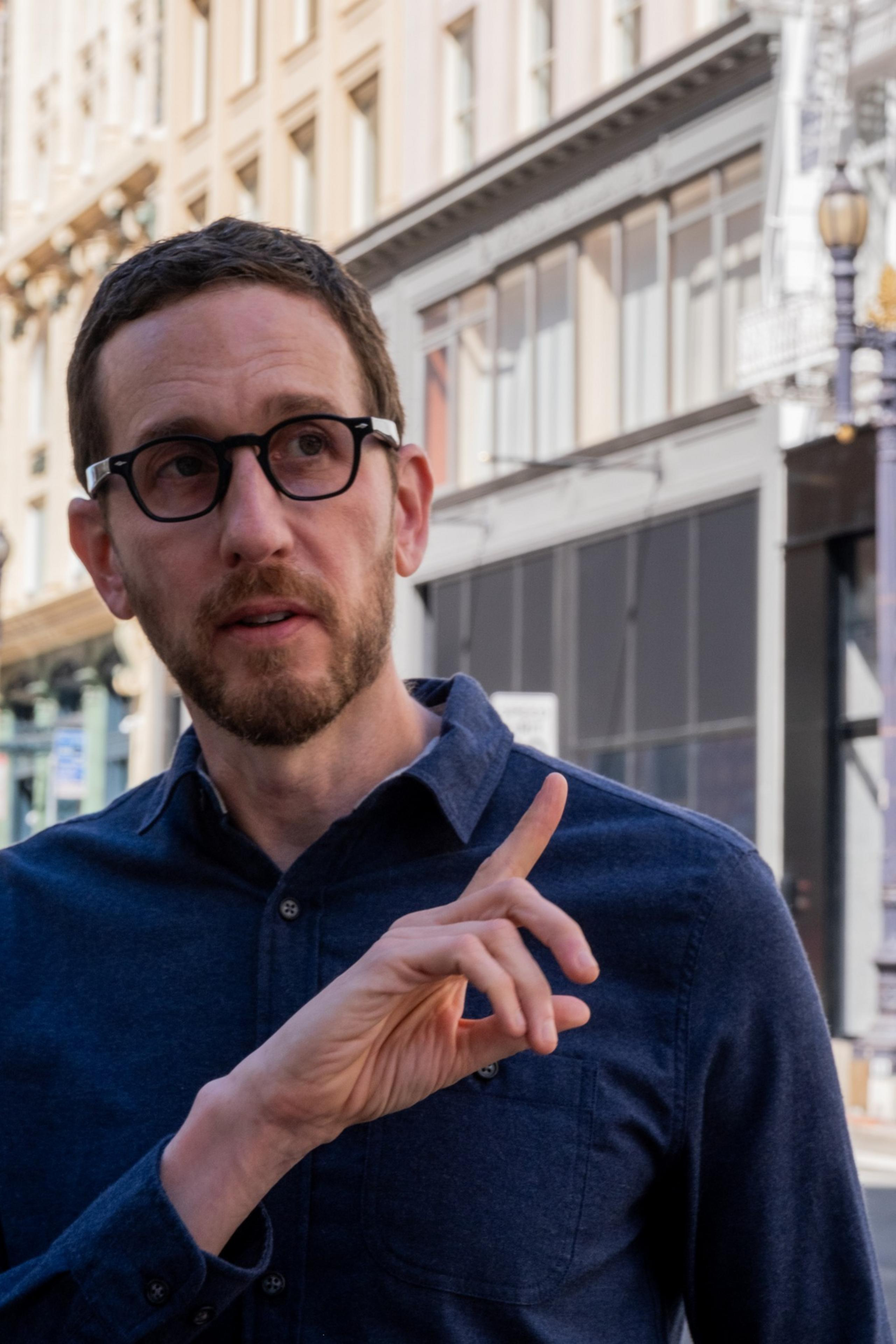 A man with short hair and glasses is pointing upwards with his right hand. He has a beard and is wearing a navy blue button-up shirt, standing in front of city buildings.