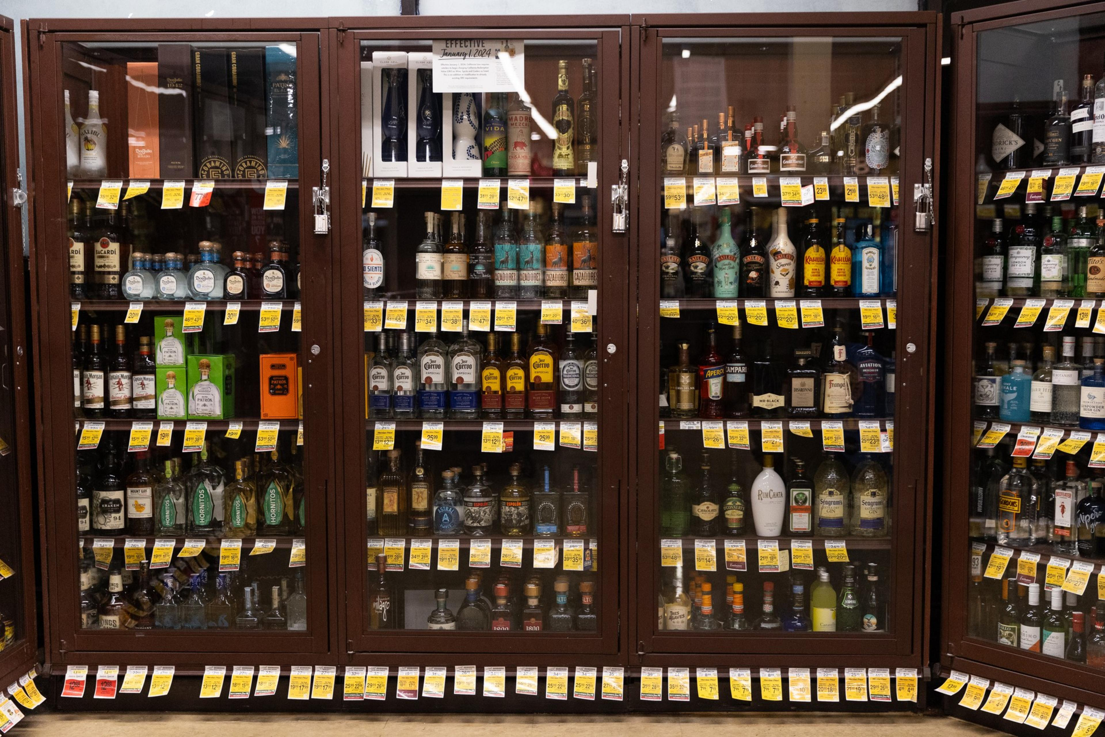 The image shows a store display of various liquor bottles in a locked cabinet, with price tags hanging on each shelf.