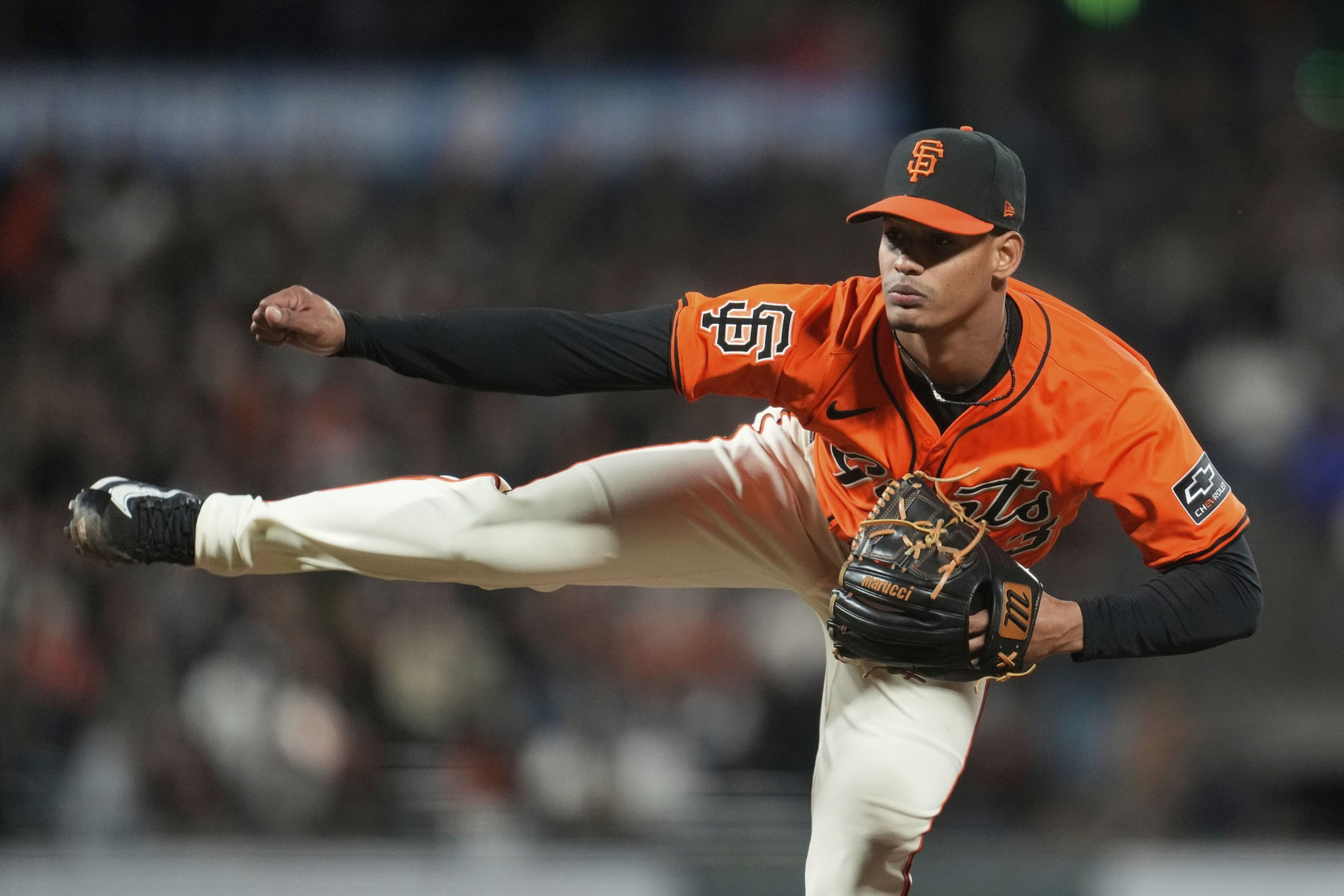 A baseball pitcher in an orange jersey with "SF" on the cap throws a pitch, with one leg high in the air. His glove is positioned near his chest.
