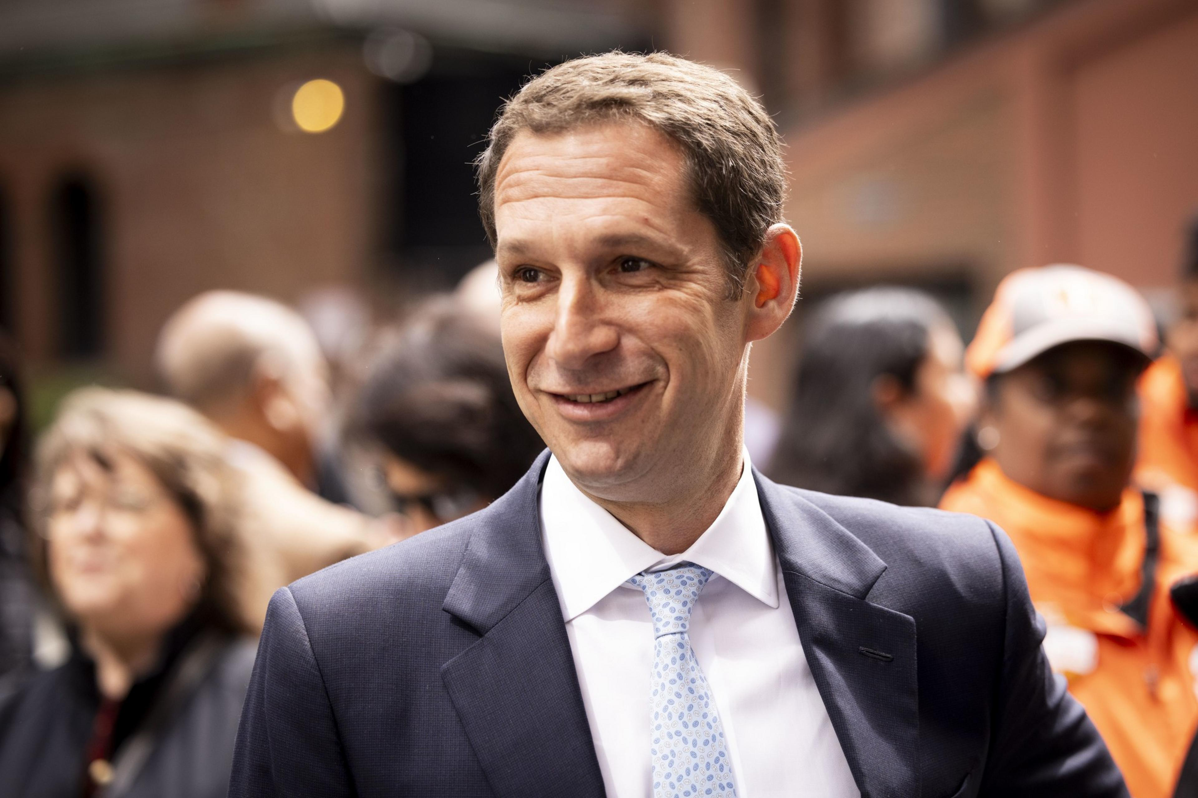 A man in a suit and light blue tie smiles outdoors, surrounded by a blurred crowd. The background includes people and brick buildings.