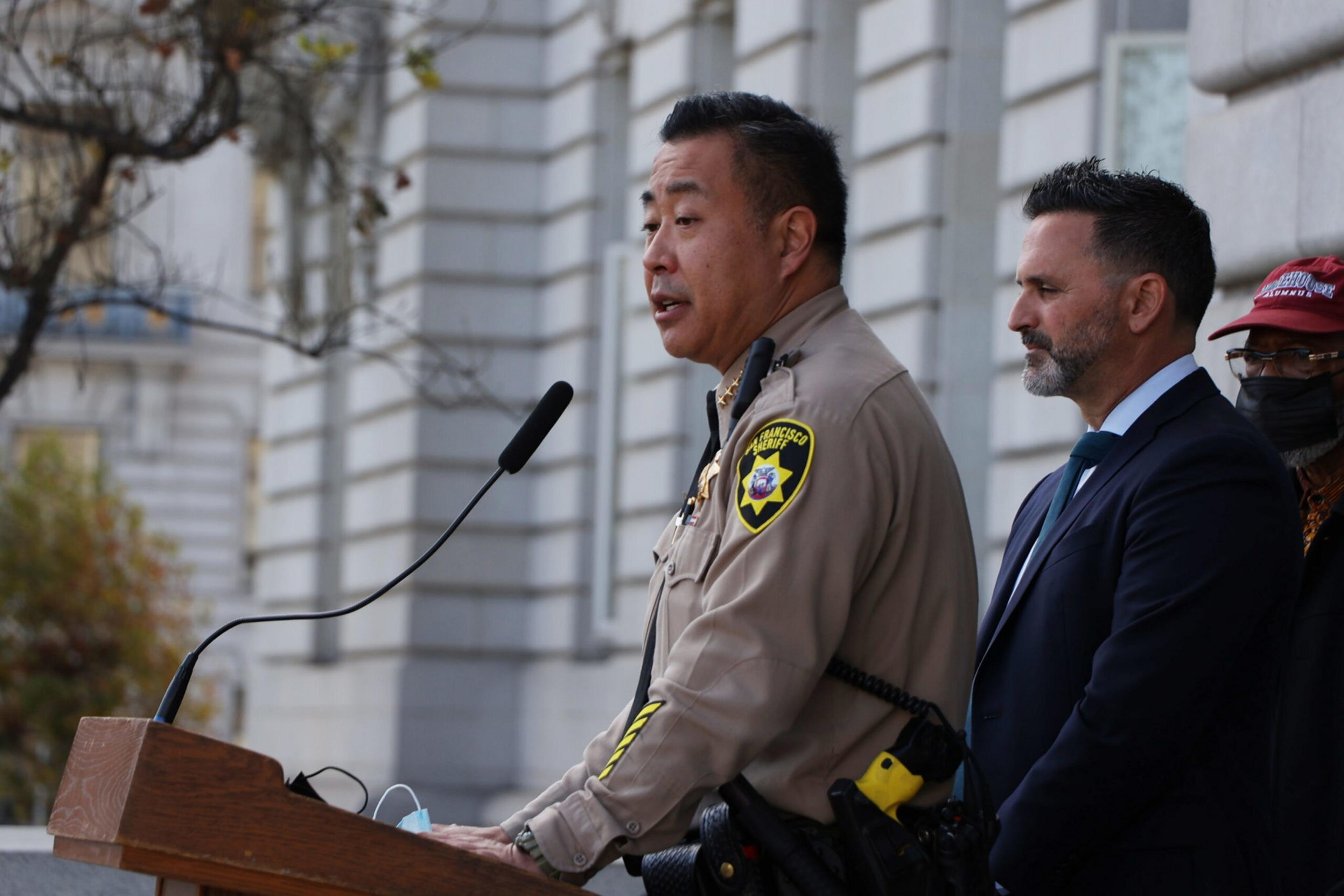 A police officer speaks at a podium with a microphone, while a man in a suit and another person in a red hat stand behind him.