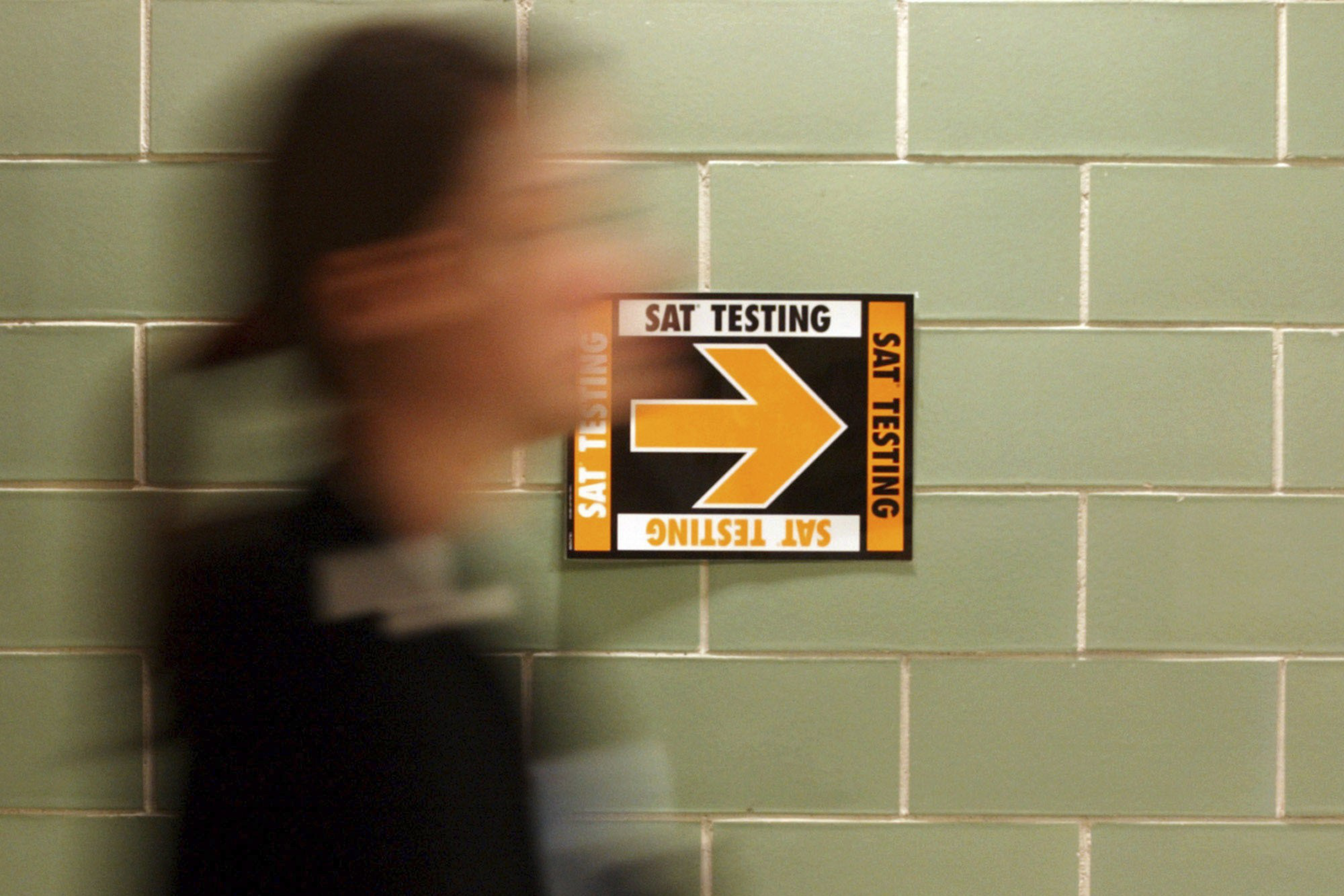 A blurred person walks past a green tiled wall with a yellow and black sign pointing right for SAT testing.