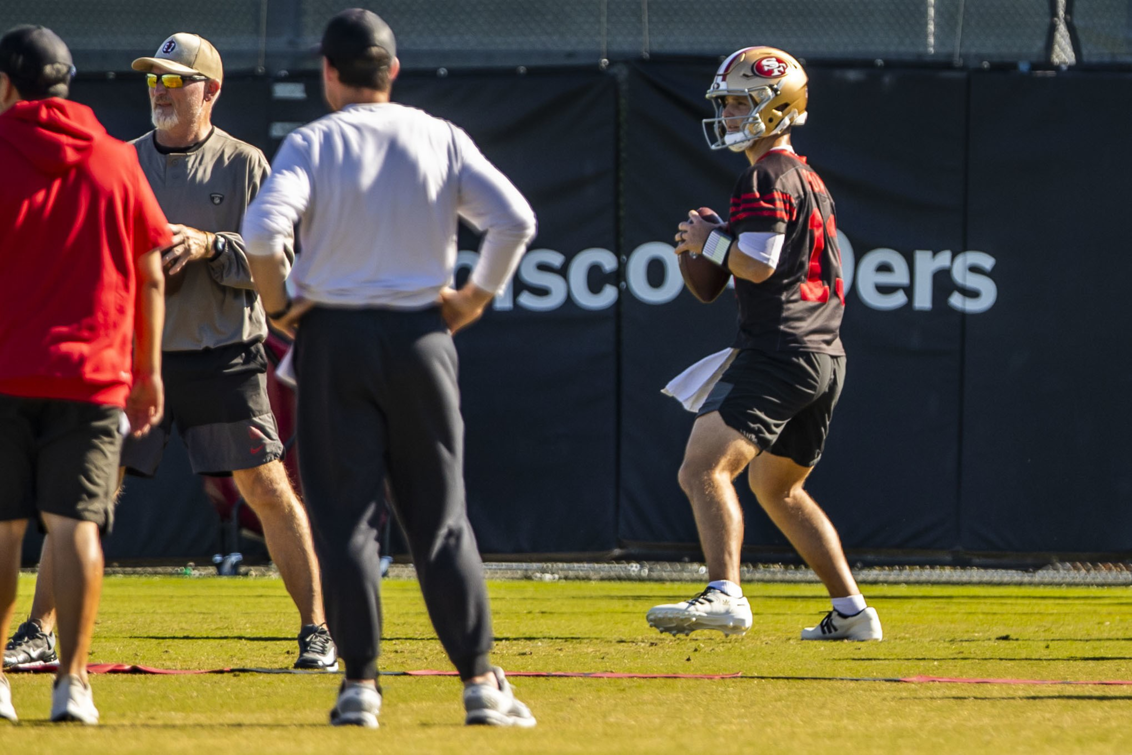A football player in a gold helmet and black uniform is holding a ball, preparing to throw, while three men in casual sportswear stand nearby on the field.