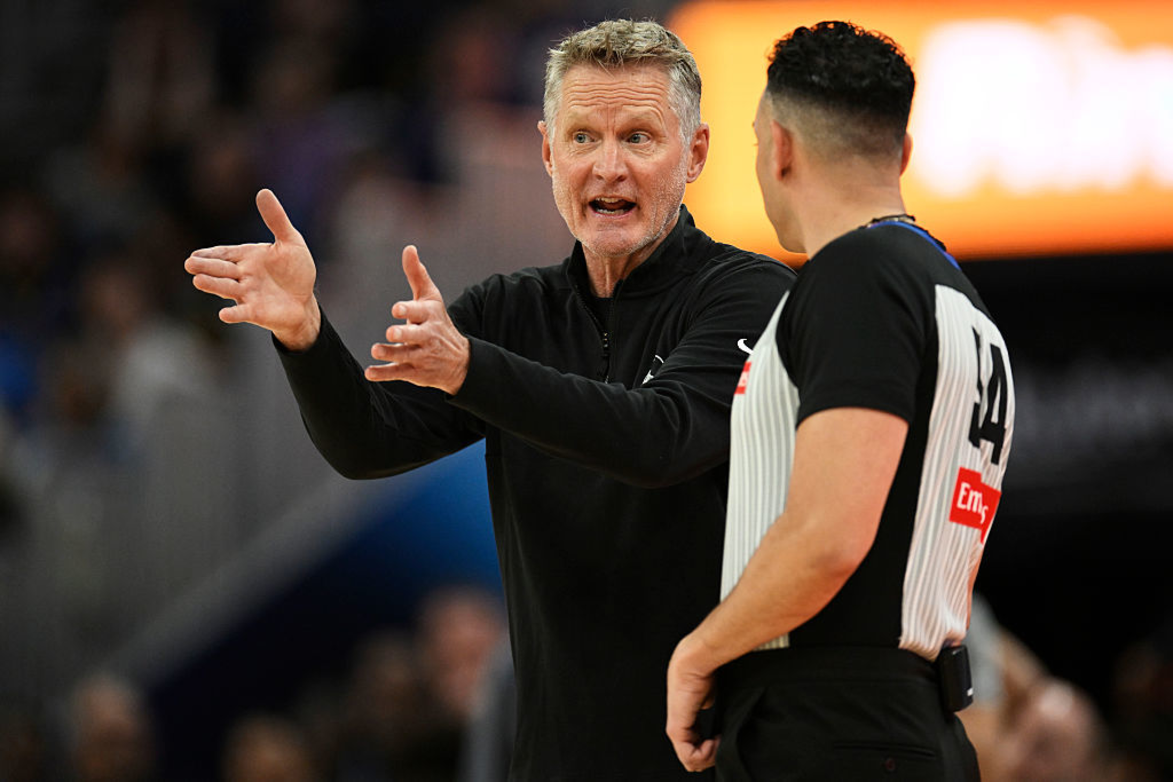 A man in a black jacket is animatedly talking to a referee in a black-and-white striped shirt, gesturing with his hands. The background is blurred.