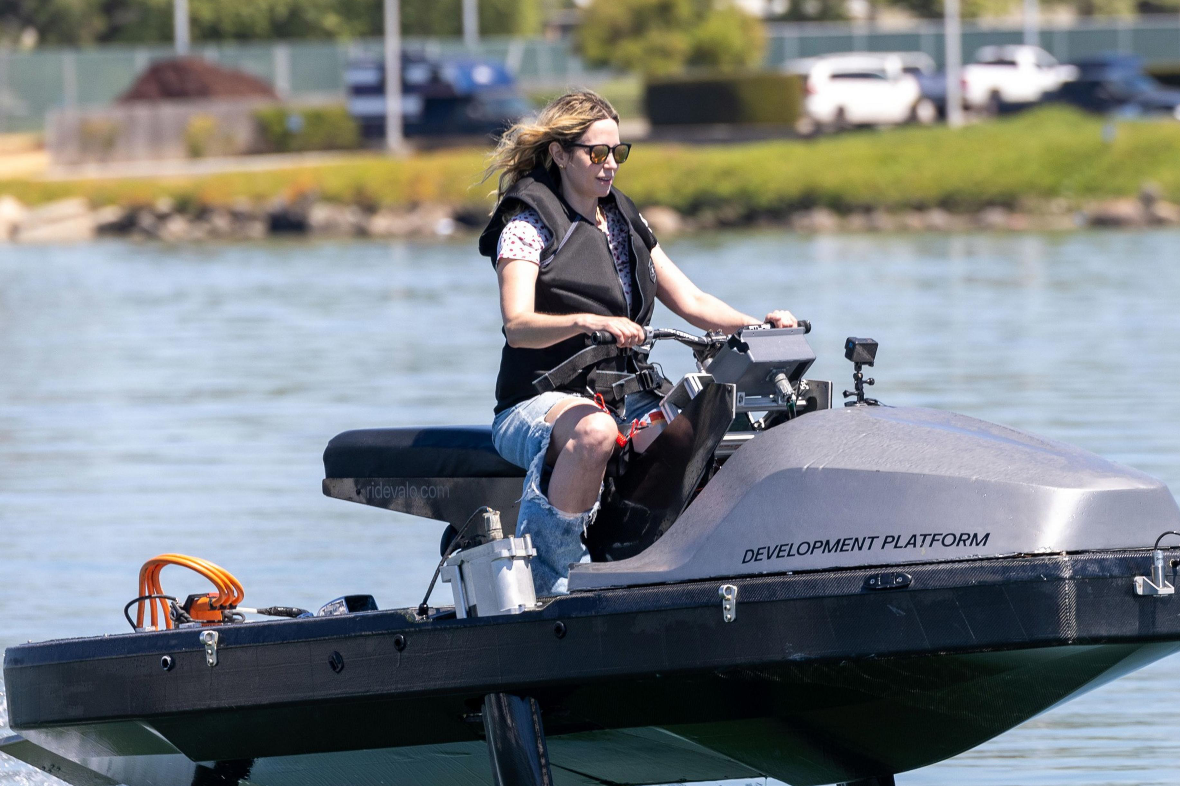 A person in sunglasses and a life jacket is riding a futuristic-looking watercraft labeled "DEVELOPMENT PLATFORM" on a calm body of water.