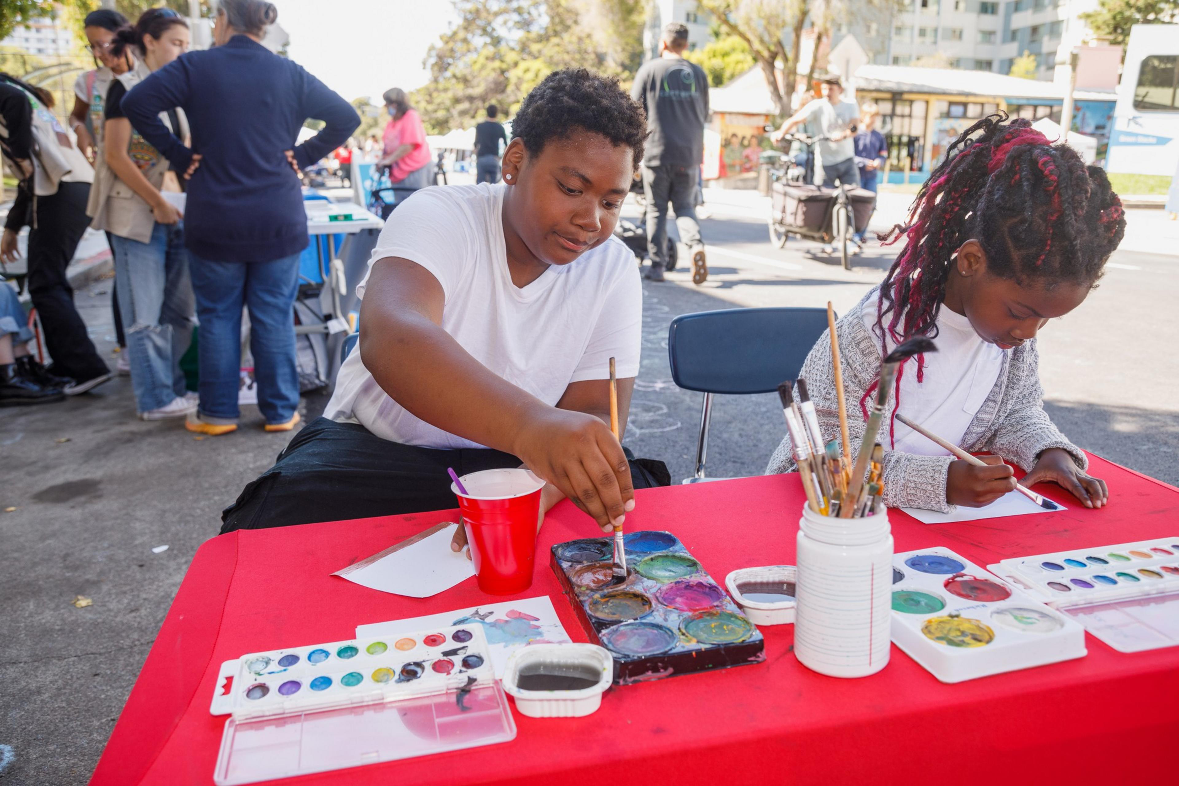 Two children are painting at a red table with art supplies, outdoors on a sunny day.