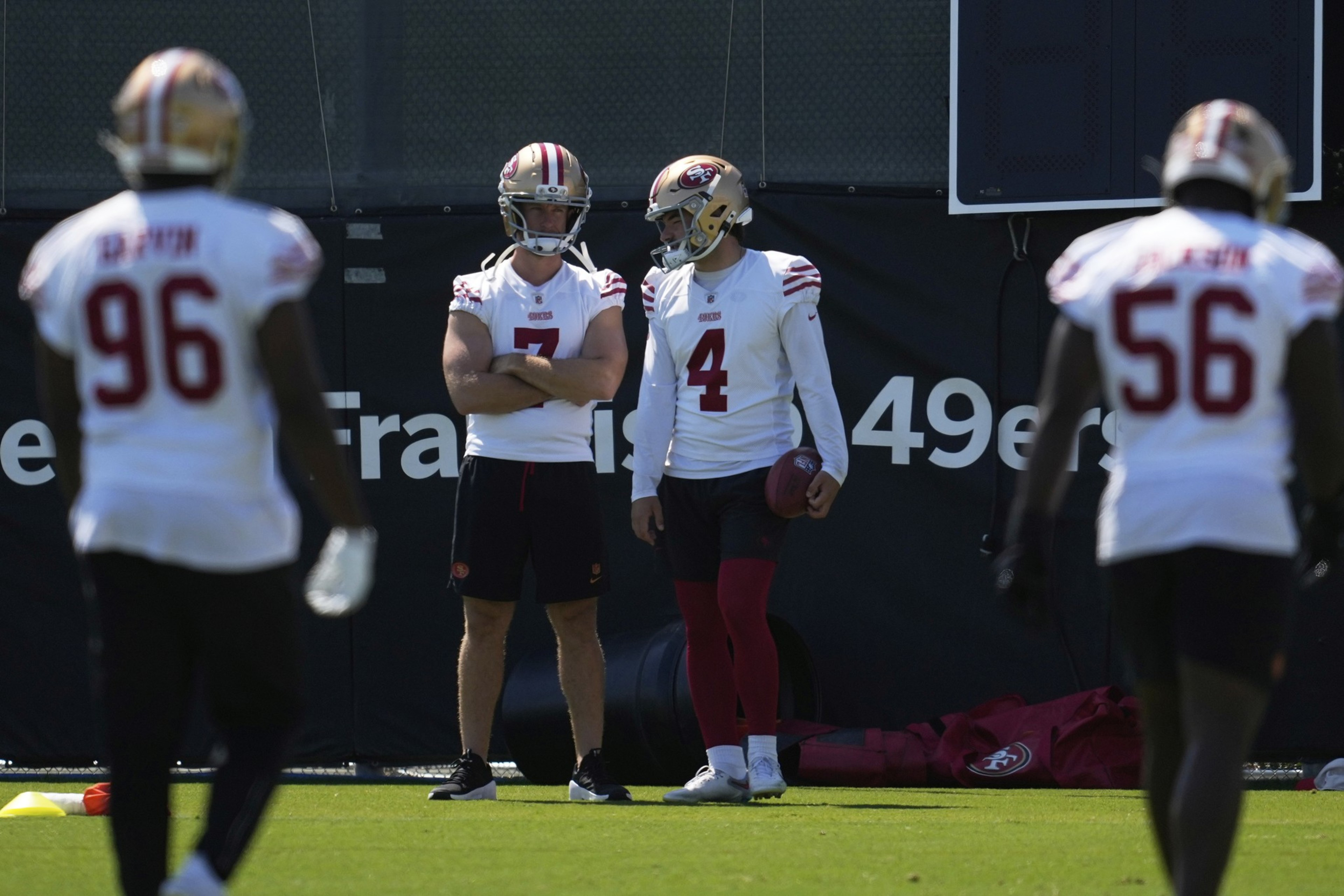 Four San Francisco 49ers players stand on a football field during practice, with two in focus talking while the others face away.