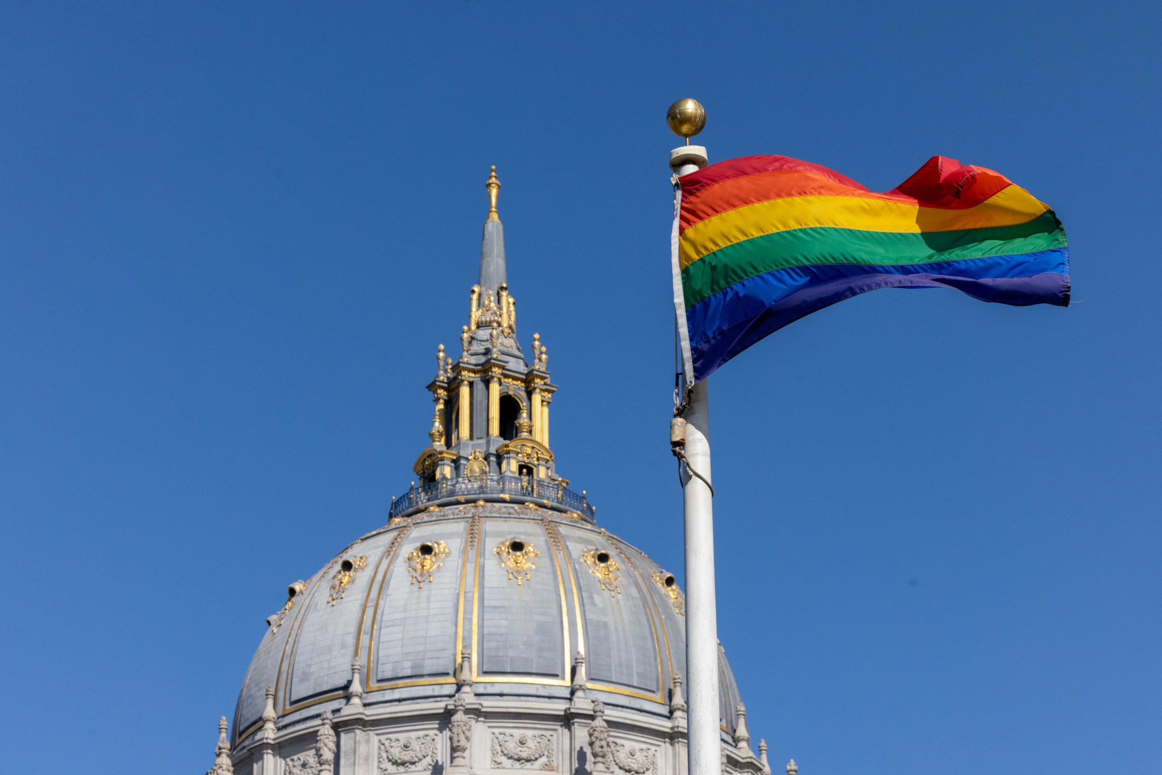 A domed building with ornate gold details stands against a clear blue sky, beside a pole with a rainbow pride flag waving.