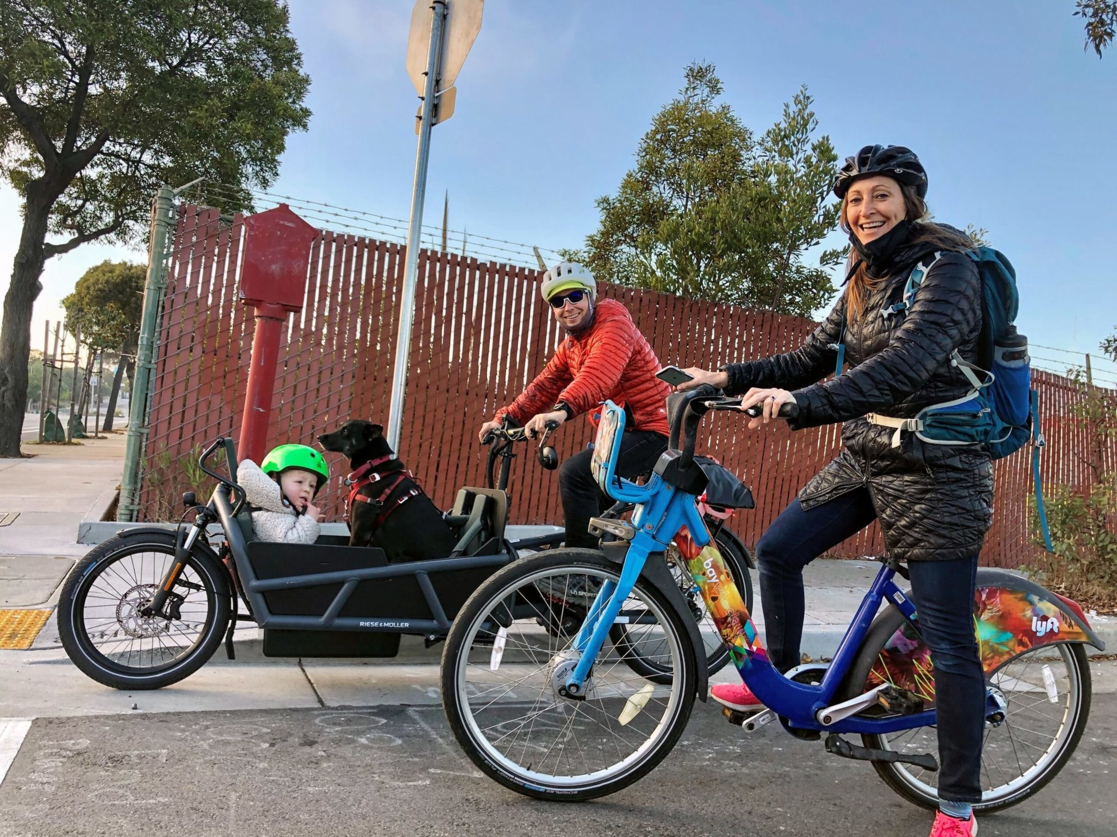 A woman and a man ride bikes; the man’s cargo bike carries a child with a green helmet and a black dog wearing a red harness.