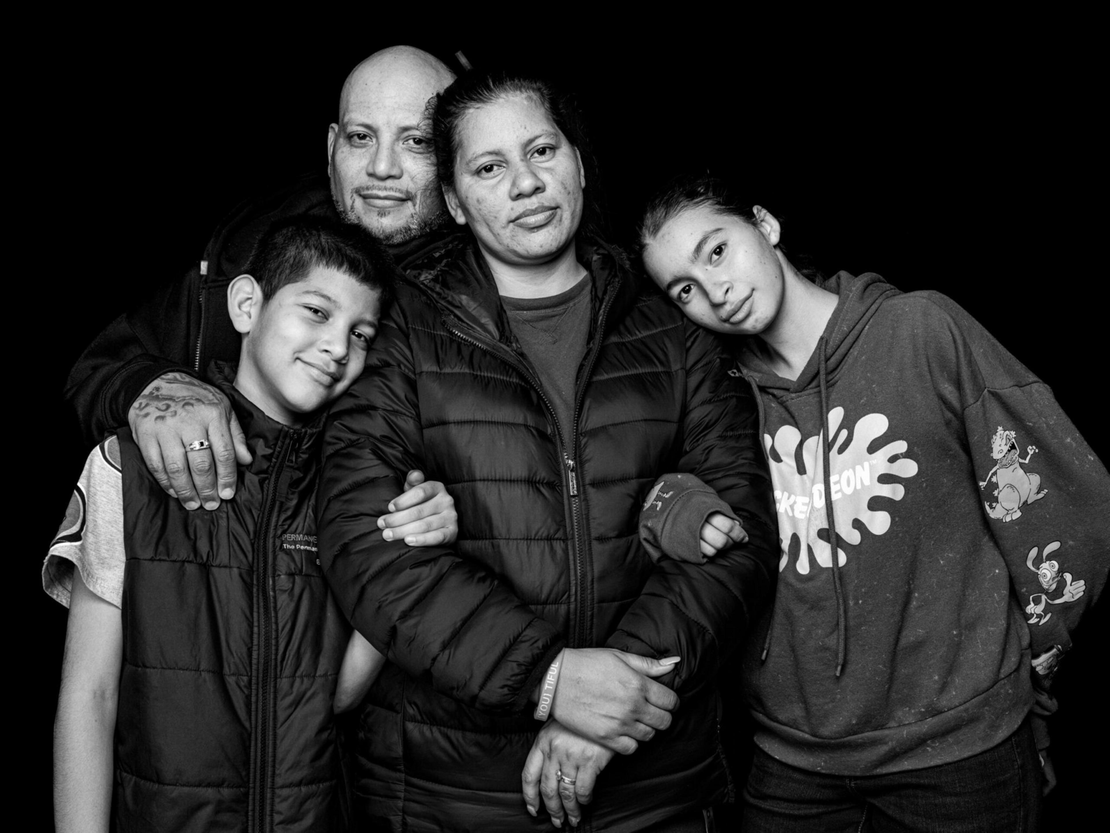A family of four stands closely together, with two children leaning on their parents, all wearing casual jackets and neutral expressions against a black background.