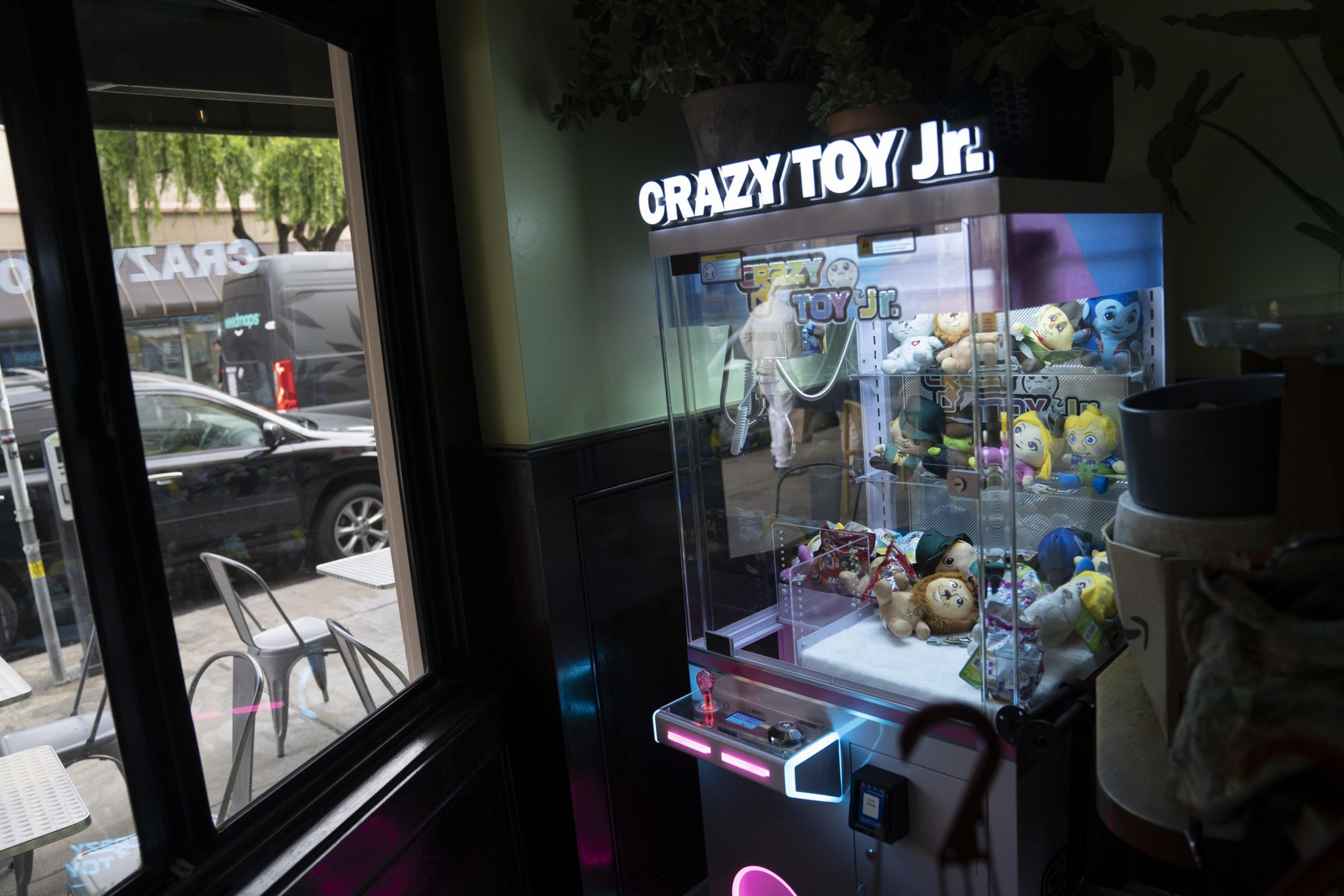 A claw machine labeled &quot;Crazy Toy Jr.&quot; filled with plush toys, including cartoon characters and a lion, is illuminated under soft lighting near potted plants.