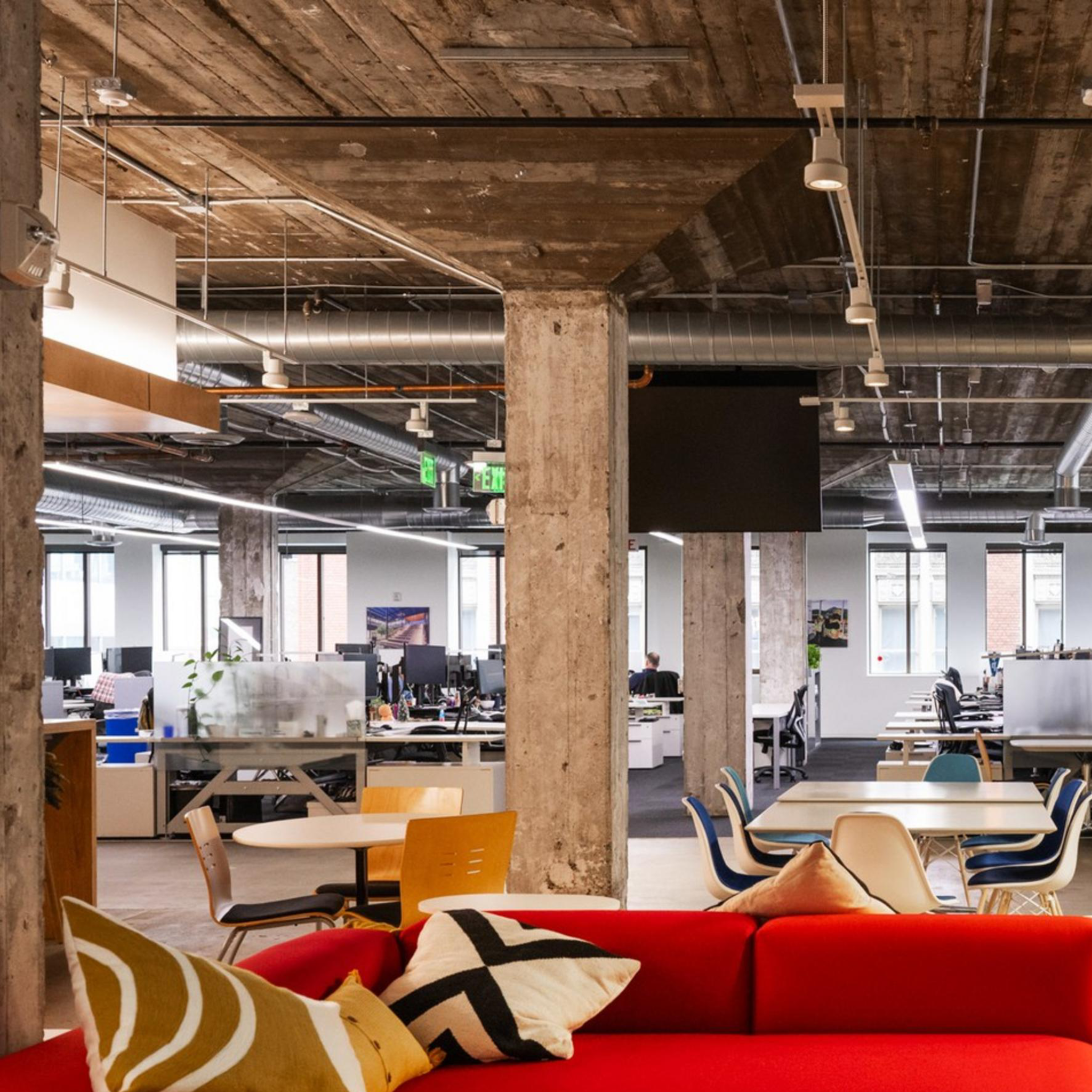 The image shows a modern office space with open seating, a red couch, a wooden reception desk, and industrial-style decor featuring exposed pipes and concrete beams.