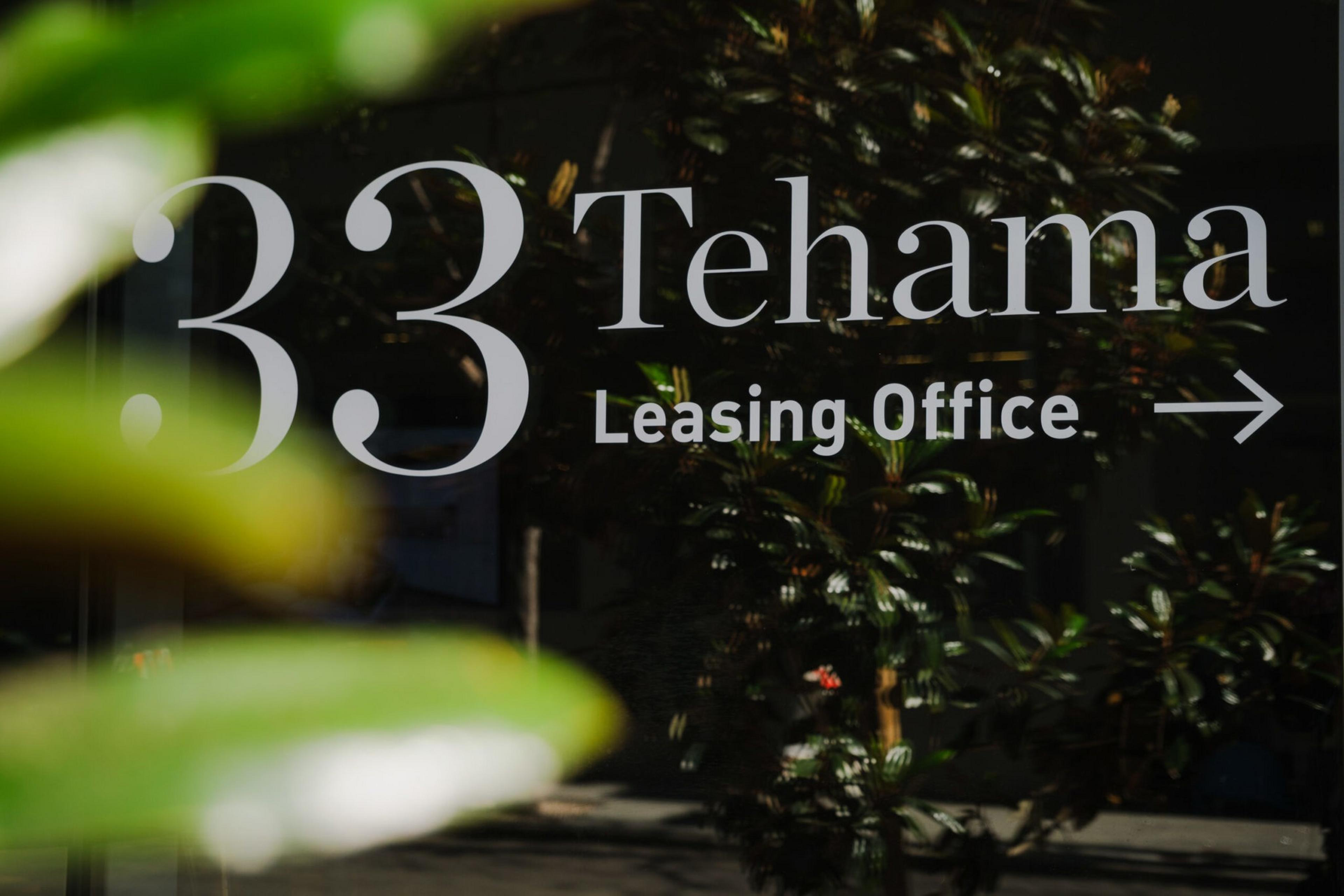 33 Tehama Leasing Office with an arrow pointing to the right is displayed on a glass window with reflections of plants and outdoor scenery.