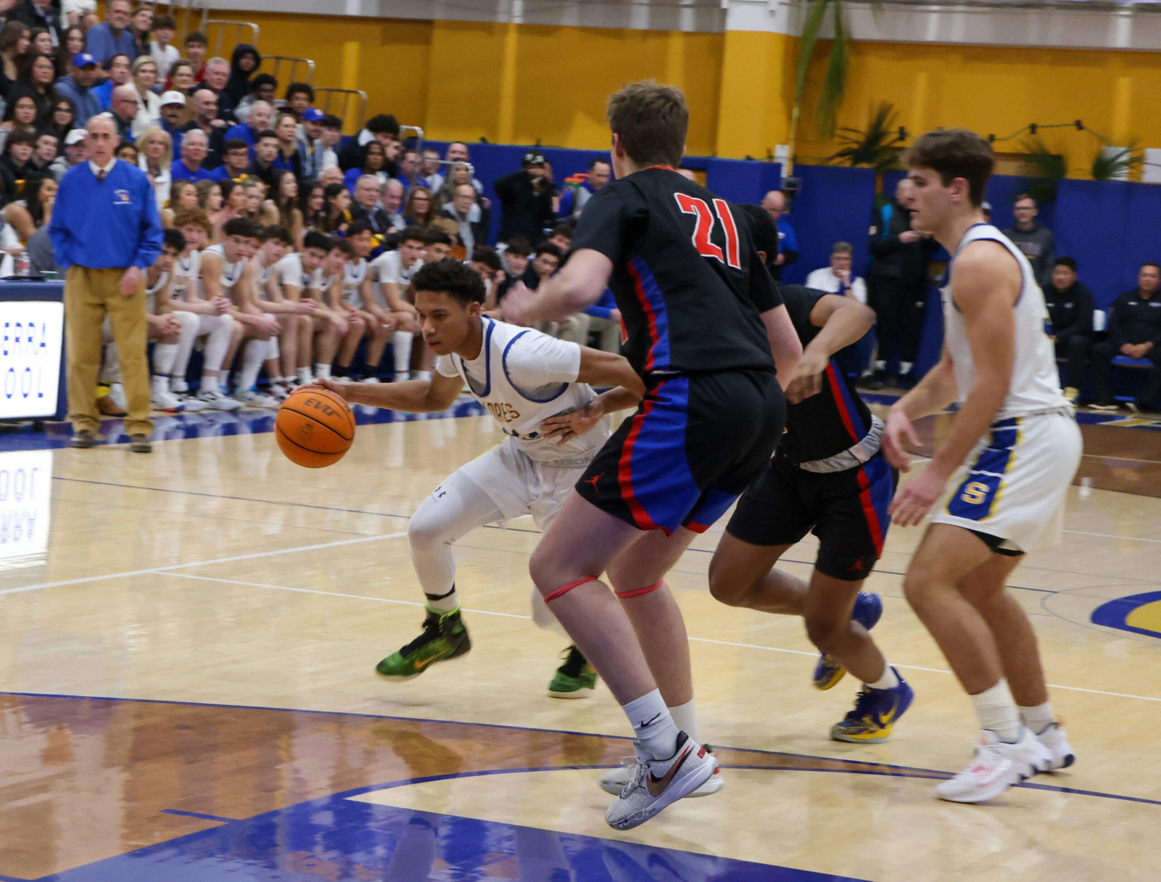 A basketball player in white dribbles past two defenders in black and red, while another player in white stands nearby, with spectators in the background.