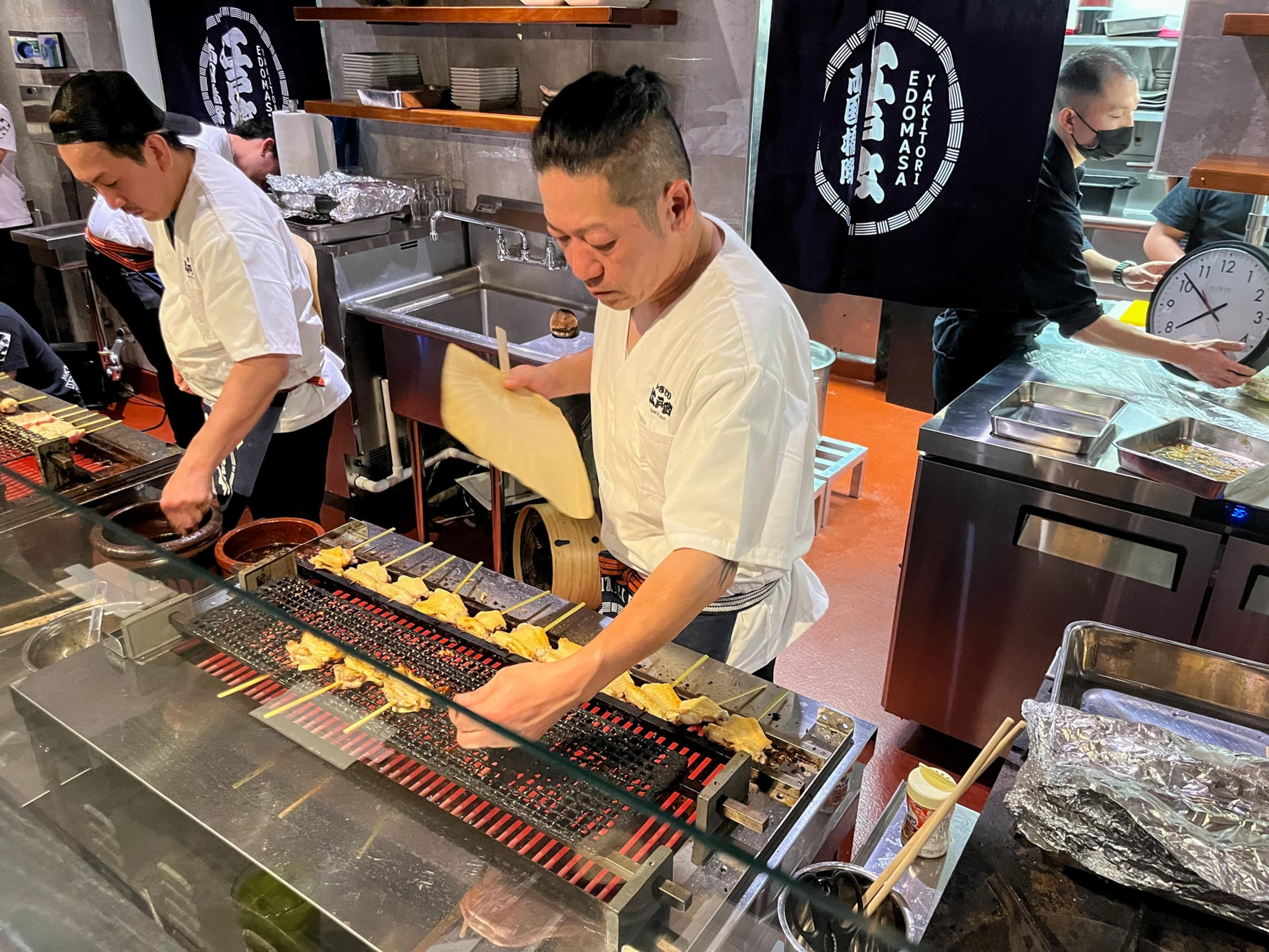 Two chefs grill skewered food over a charcoal grill in a busy kitchen, while another person in the background handles a large clock.
