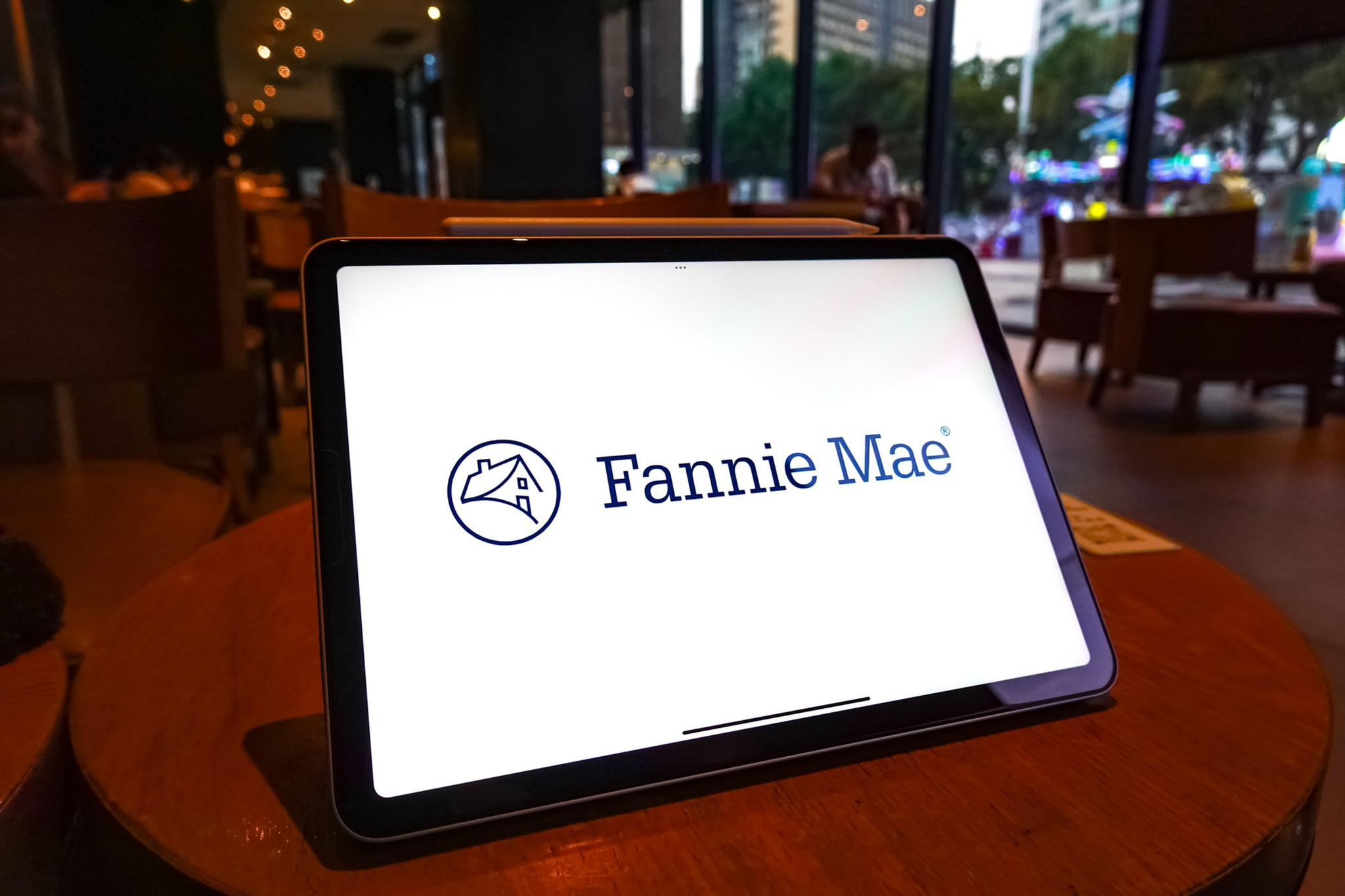 A tablet on a wooden table displays the Fannie Mae logo in a dimly lit indoor café with large windows showing city lights outside.