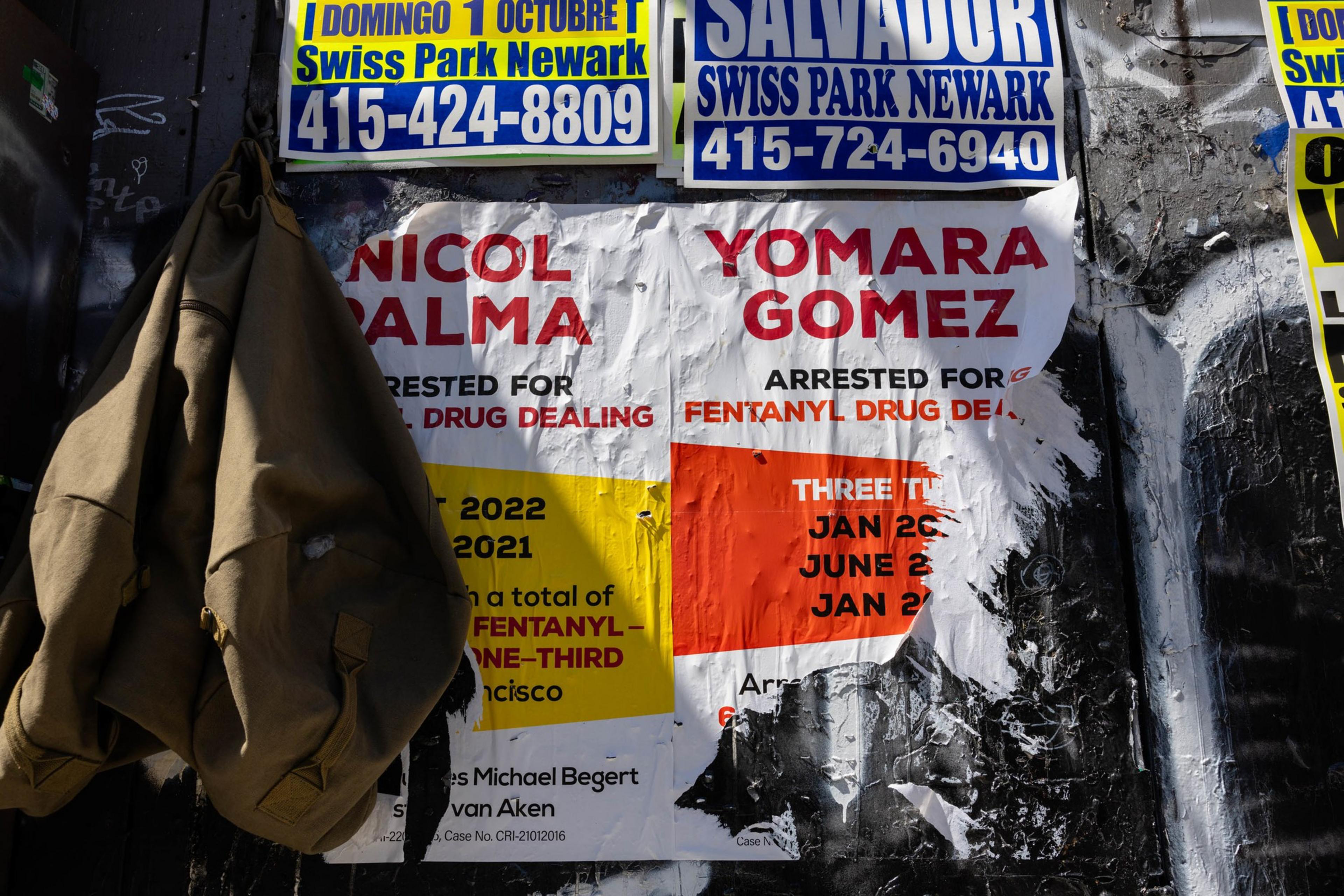 Layered posters on a wall announce arrests, with a tattered jacket hanging to the side.