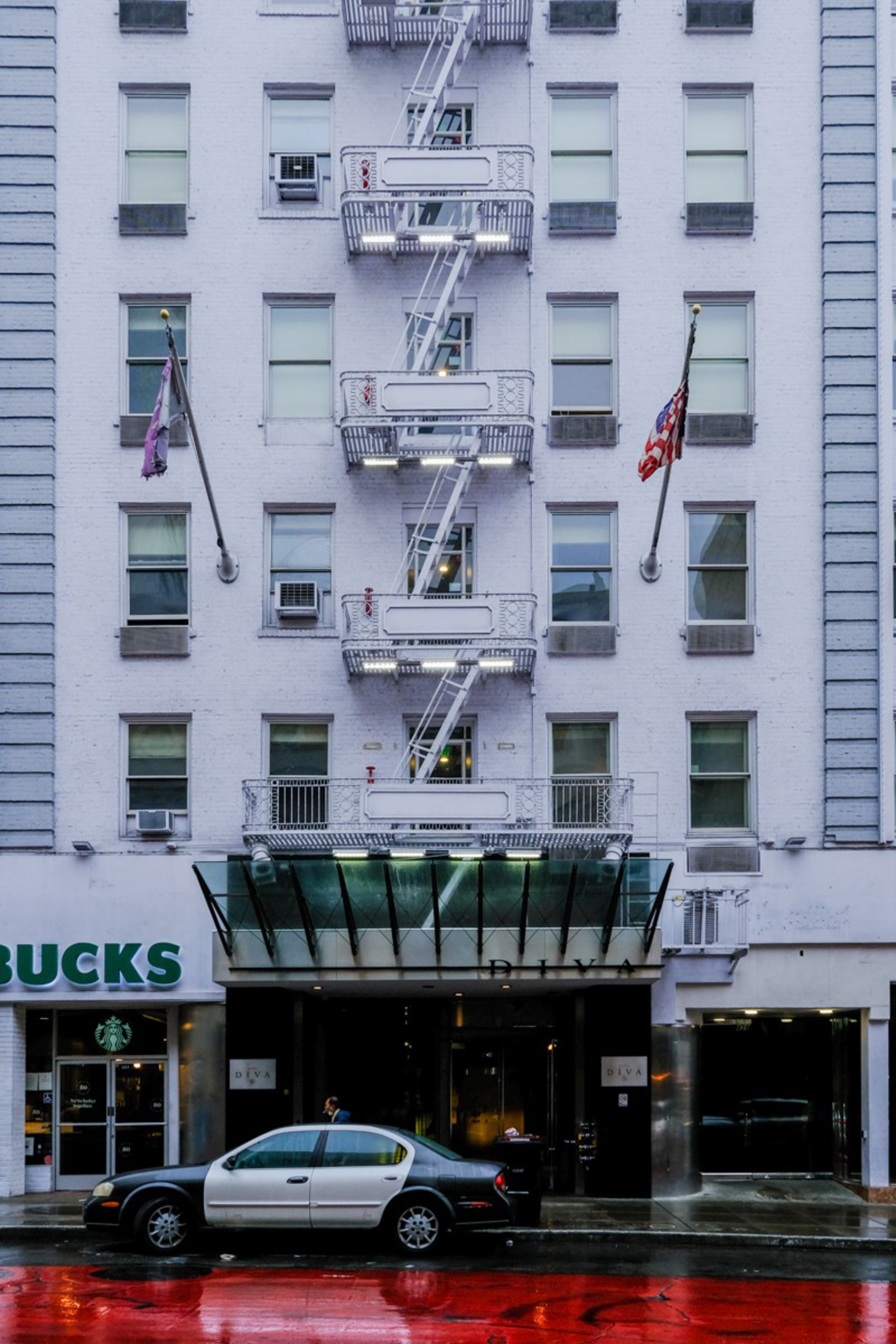 A white multi-story building has fire escapes, an American flag, and a Starbucks storefront, with a black-and-white car parked on a wet red street.