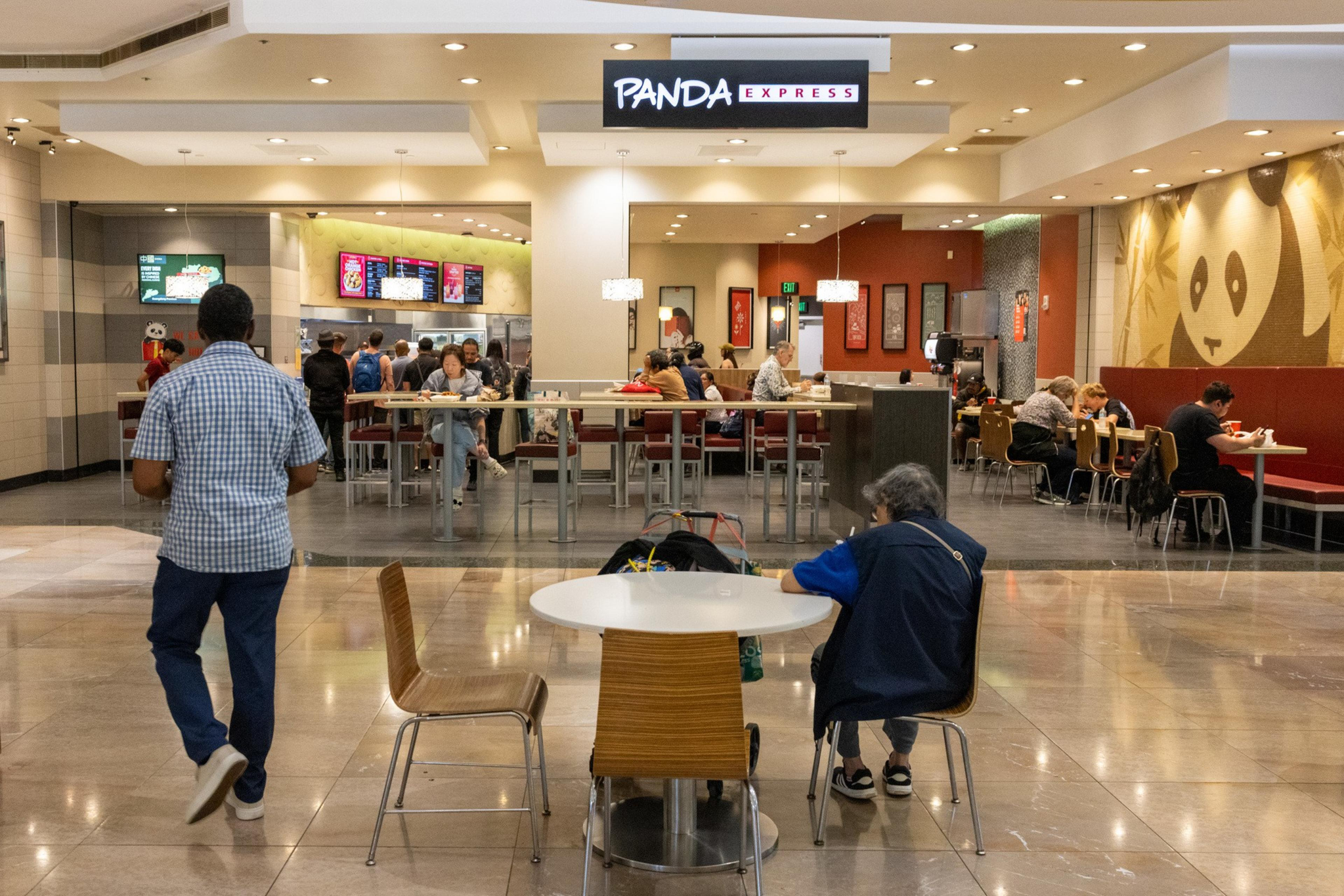 A Panda Express restaurant in a mall food court with customers seated and standing in line, and a large panda logo on the wall to the right.