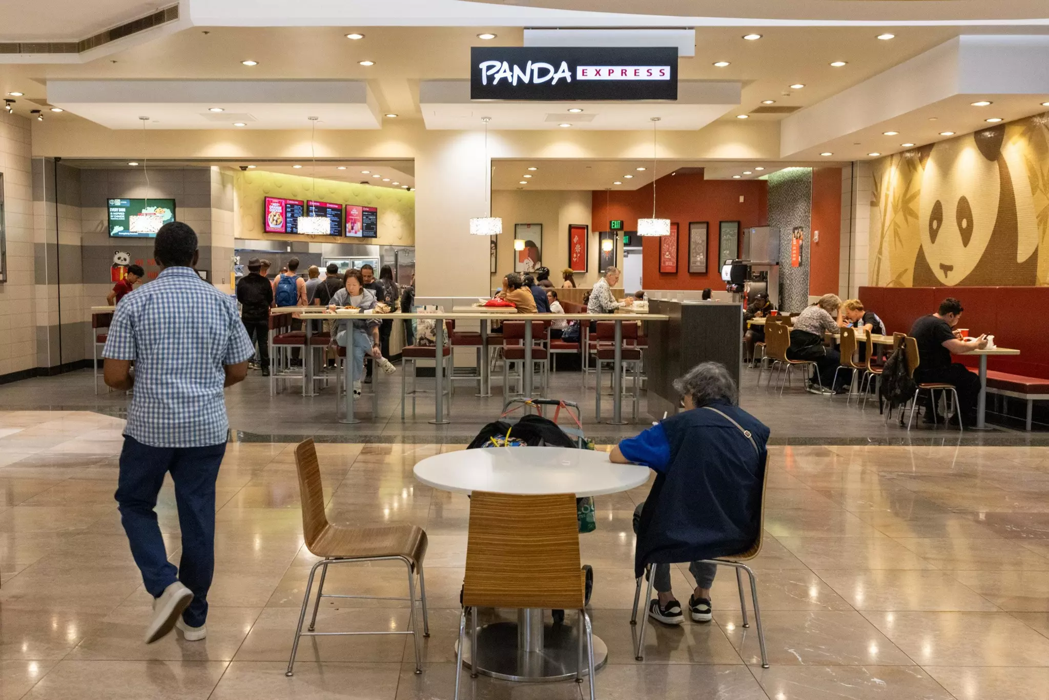 A Panda Express restaurant in a mall food court with customers seated and standing in line, and a large panda logo on the wall to the right.