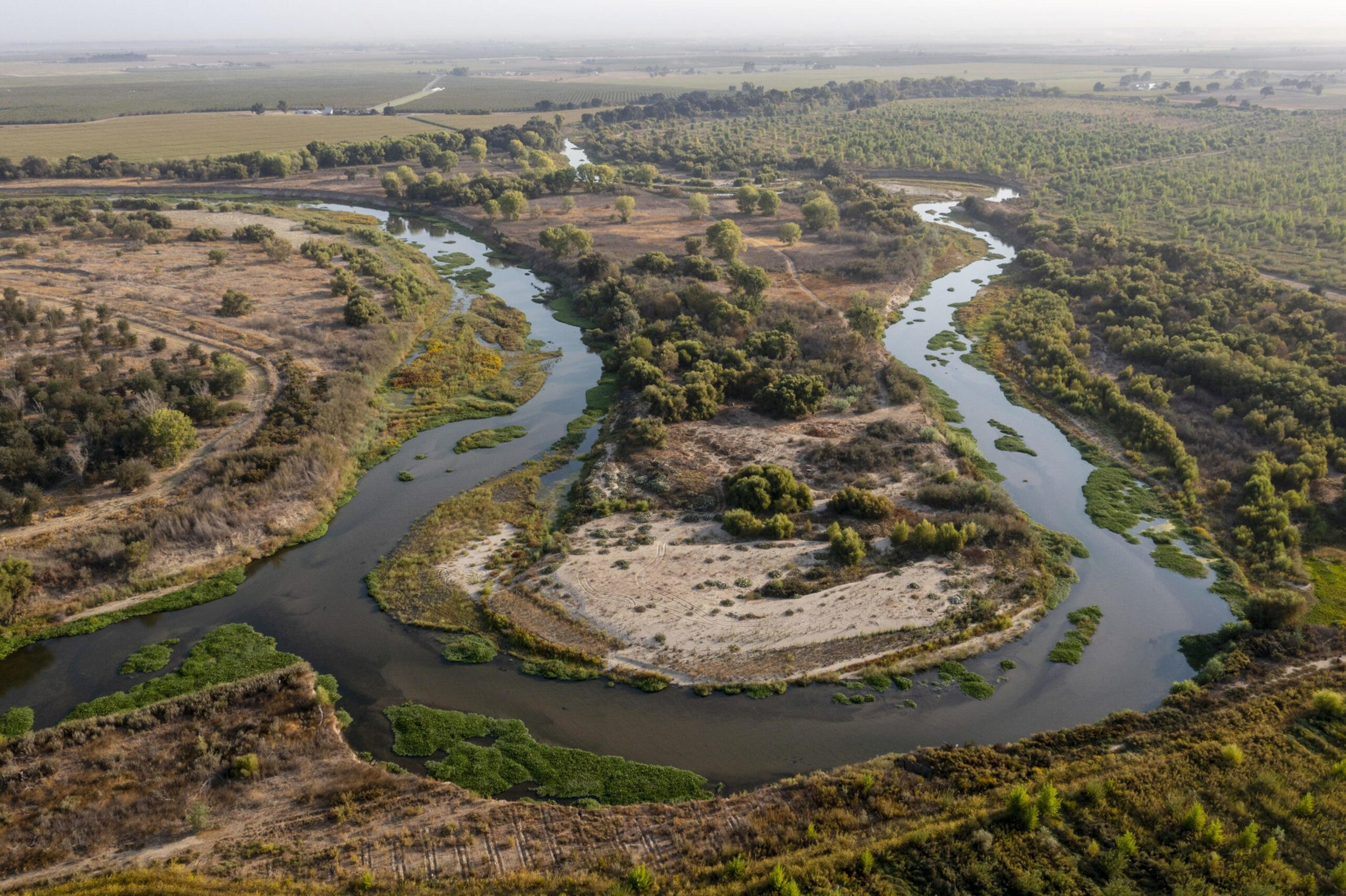 A winding river flows through a dry, patchy landscape dotted with green shrubs and trees, surrounded by open fields and farmland.
