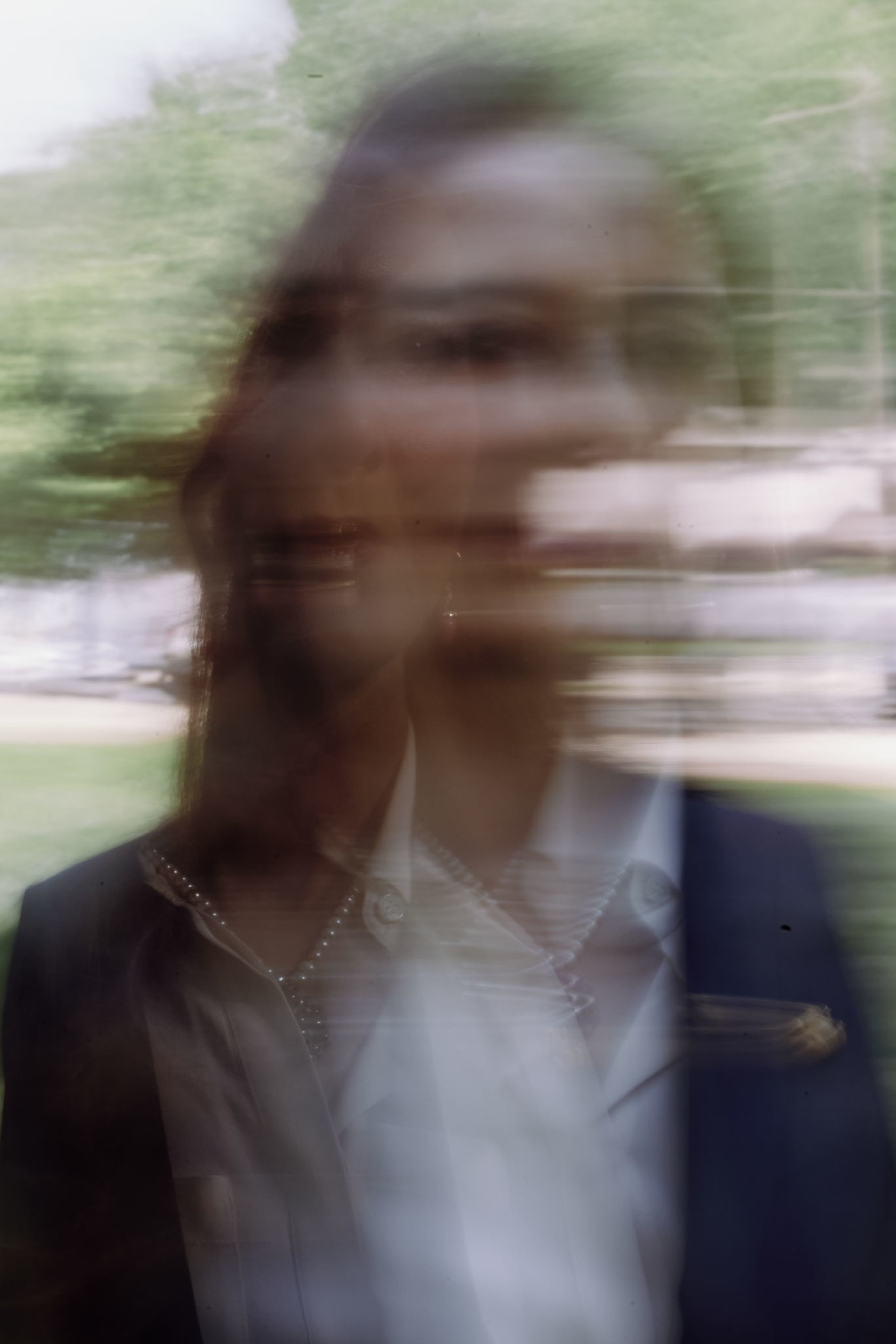 The image shows a blurred, double-exposed portrait of a person outdoors, with greenery in the background, giving a ghostly, distorted effect.