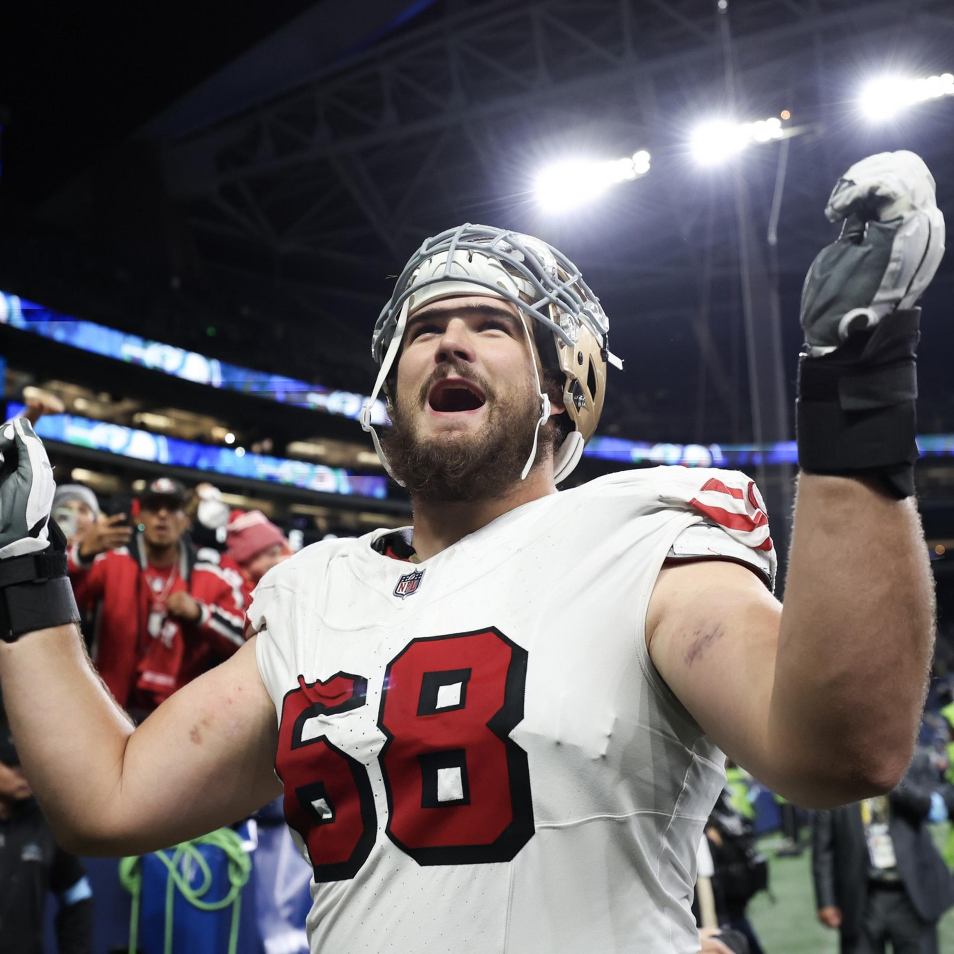 A football player wearing a white jersey with number 68 raises his arms and cheers on the field while fans celebrate behind him.
