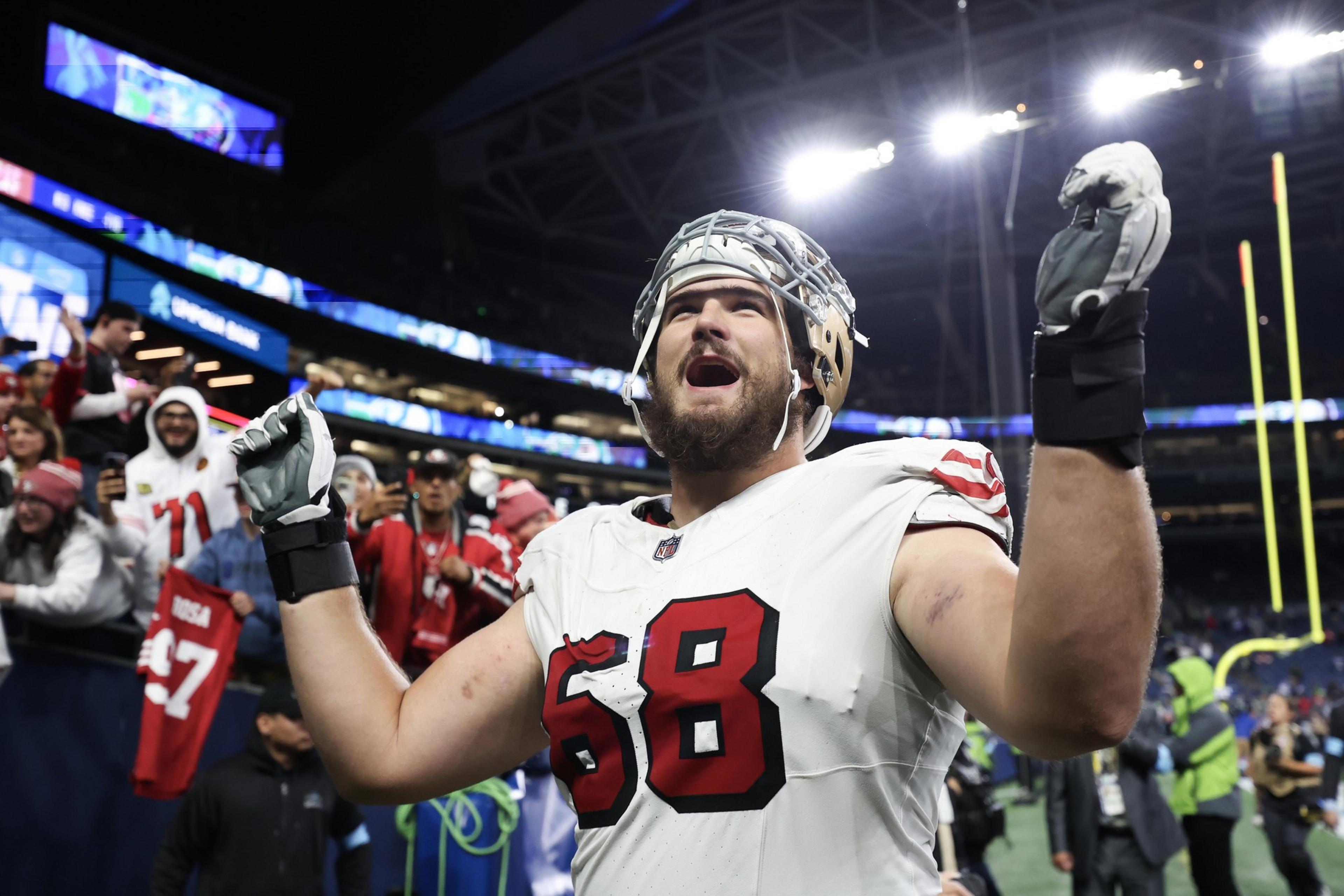 A football player wearing a white jersey with number 68 raises his arms and cheers on the field while fans celebrate behind him.
