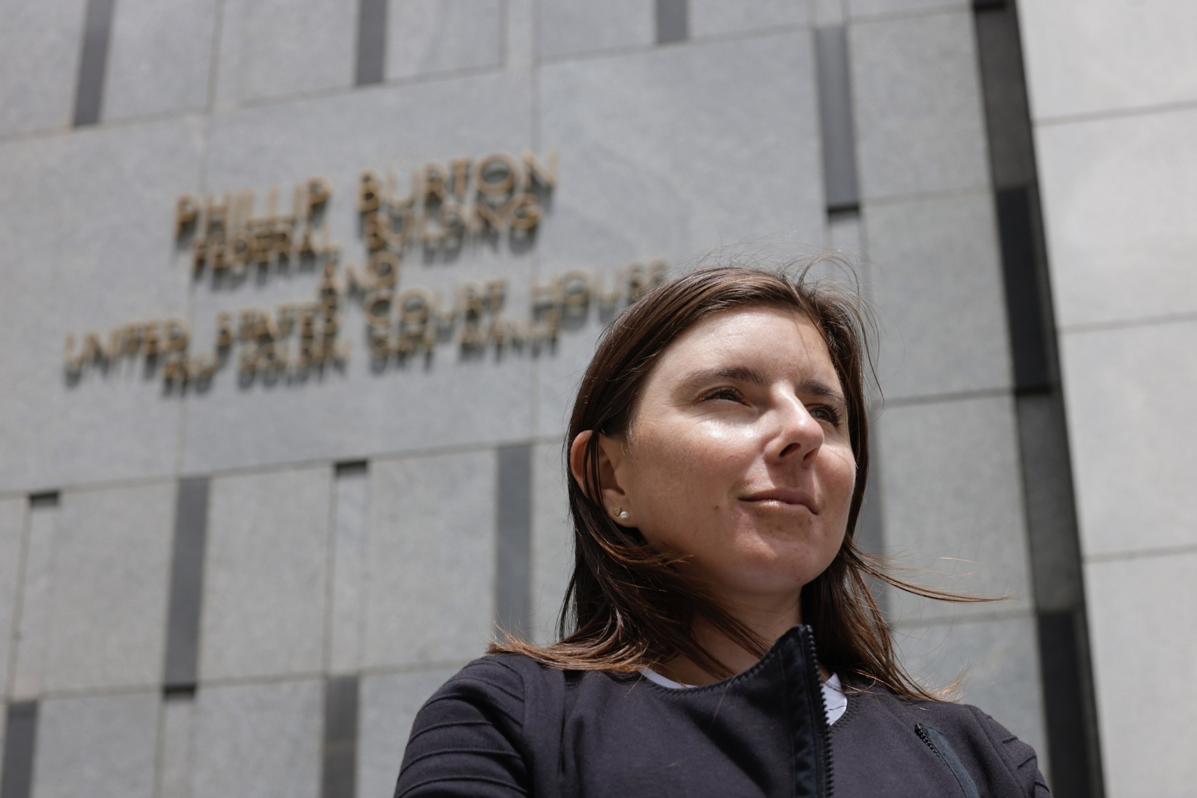 A woman stands in front of a gray building with the sign &quot;PHILLIP BURTON FEDERAL BUILDING AND UNITED STATES COURT HOUSE&quot; visible in the background.