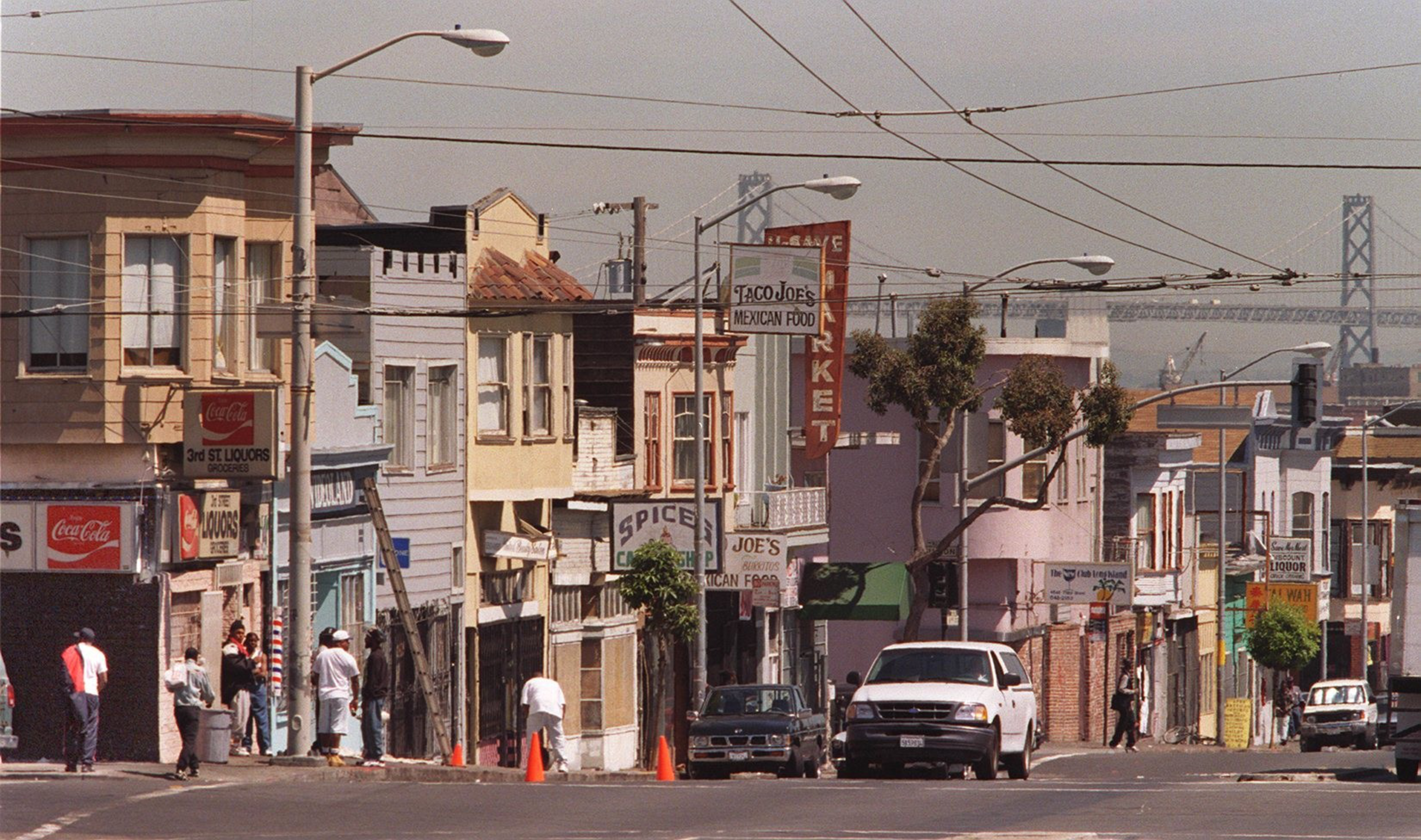 The image shows a city street lined with small shops, colorful old buildings, and parked vehicles. People walk and gather near store entrances. A bridge is visible in the background.