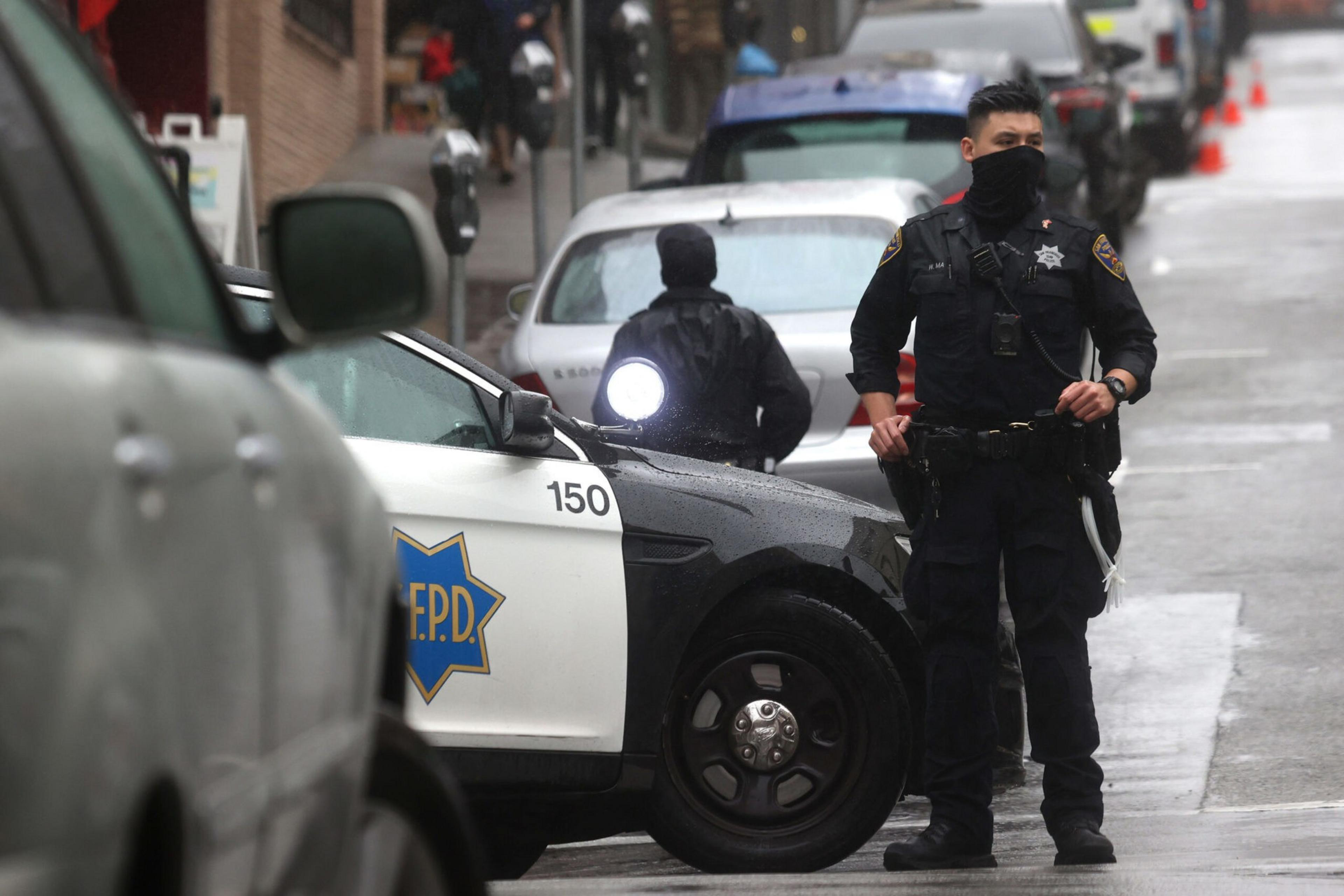A police officer in black uniform and face covering stands near a black-and-white police car blocking a rainy street with parked cars.