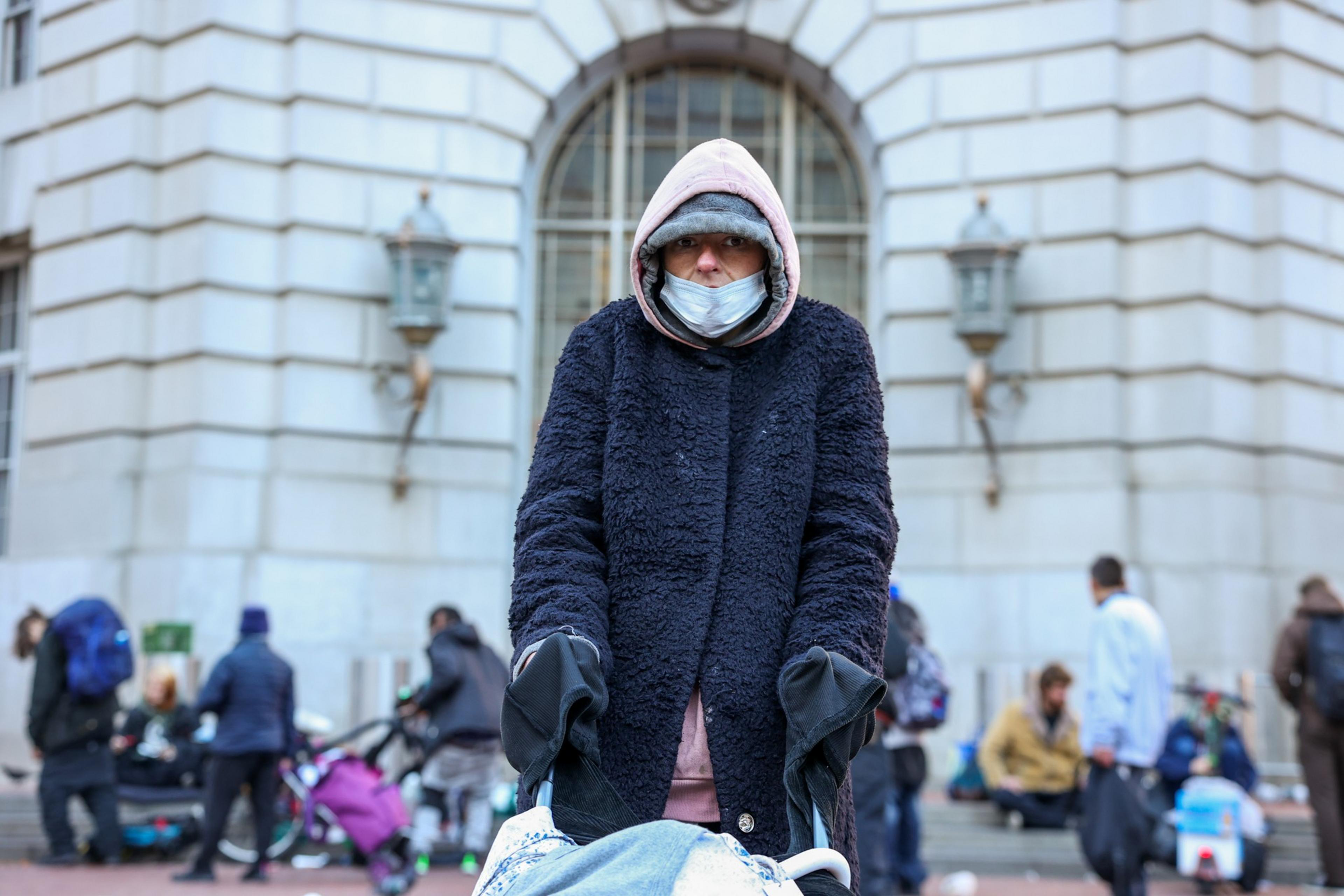 A person bundled in a heavy coat, gloves, and hood wears a mask while pushing a cart, with people and a building in the blurred background.