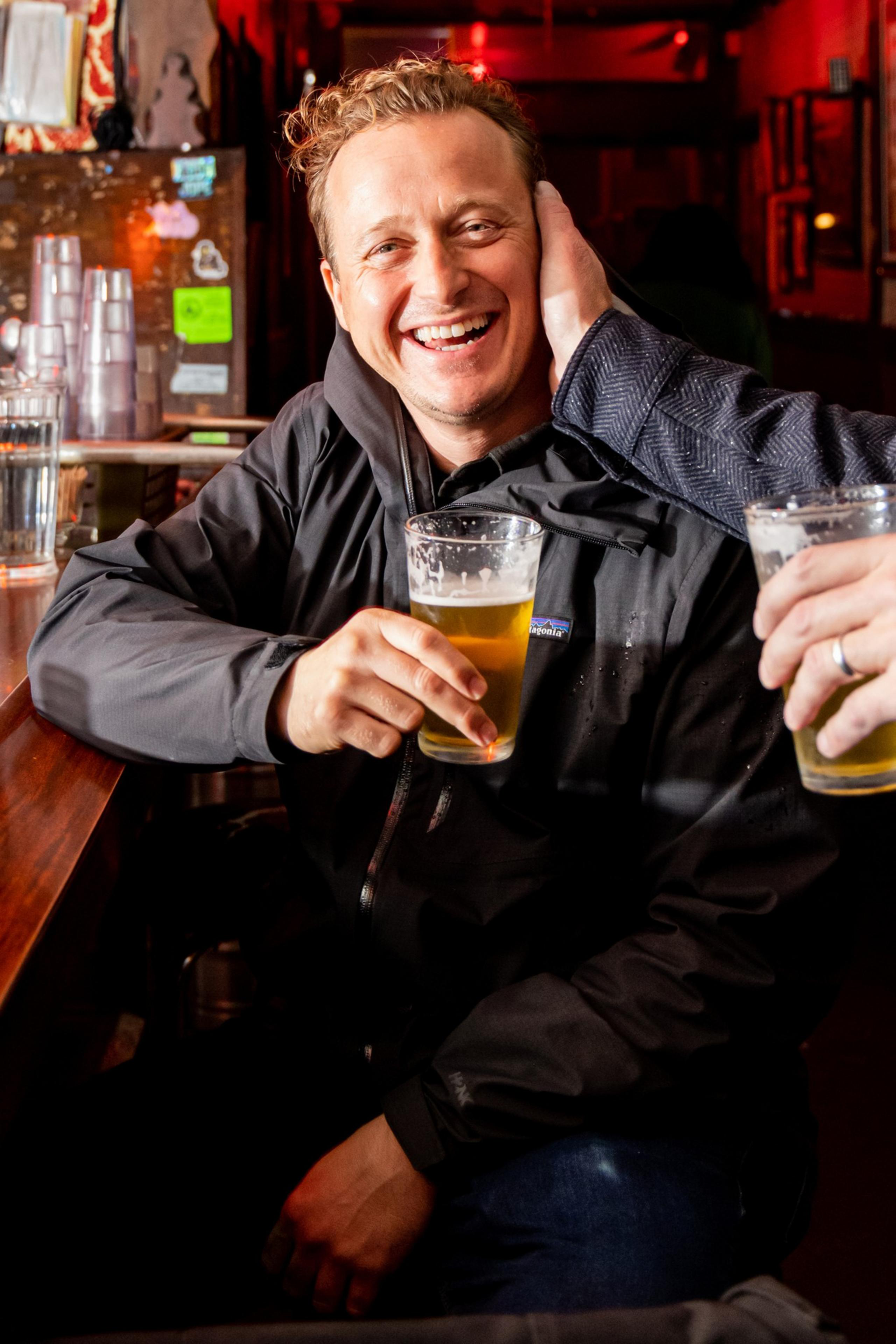 A smiling man sits at a bar, holding a pint of beer. He's wearing a black jacket, and another person playfully touches his head, also holding a beer.